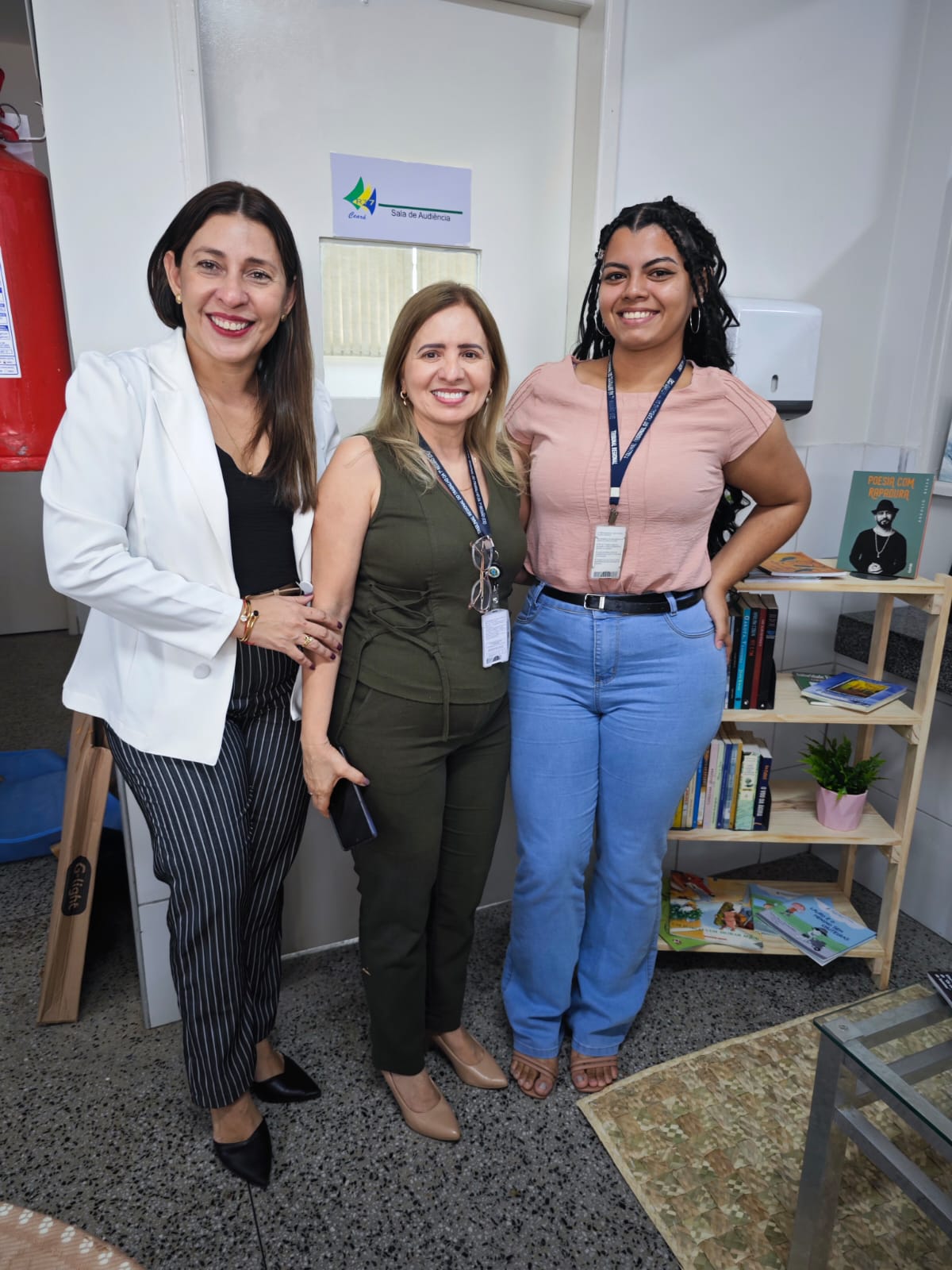 Foto da Juíza do trabalho Rossana Talia, bibliotecária Rejane Façanha e ex estagiária Eulália. Elas estão em pé, sorridentes e em frente ao ponto de leitura com estante de livros