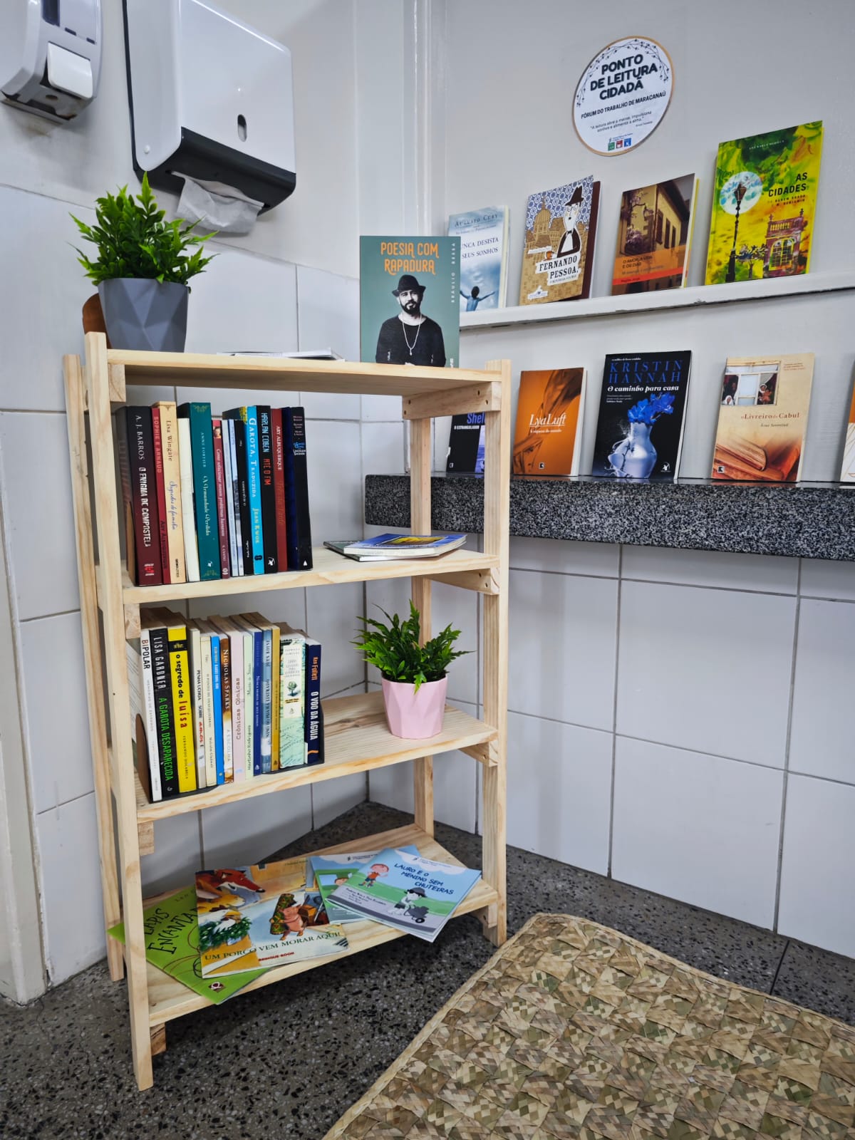 Foto do ponto de leitura. Tem estante com livros, jarro e há obras colocadas em prateleiras na parede