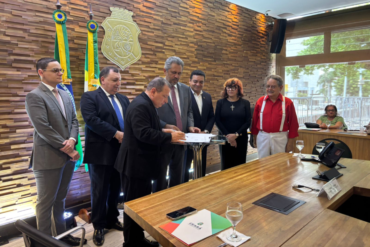 Homens de paletó ao redor de uma mesa, um deles assina um papel