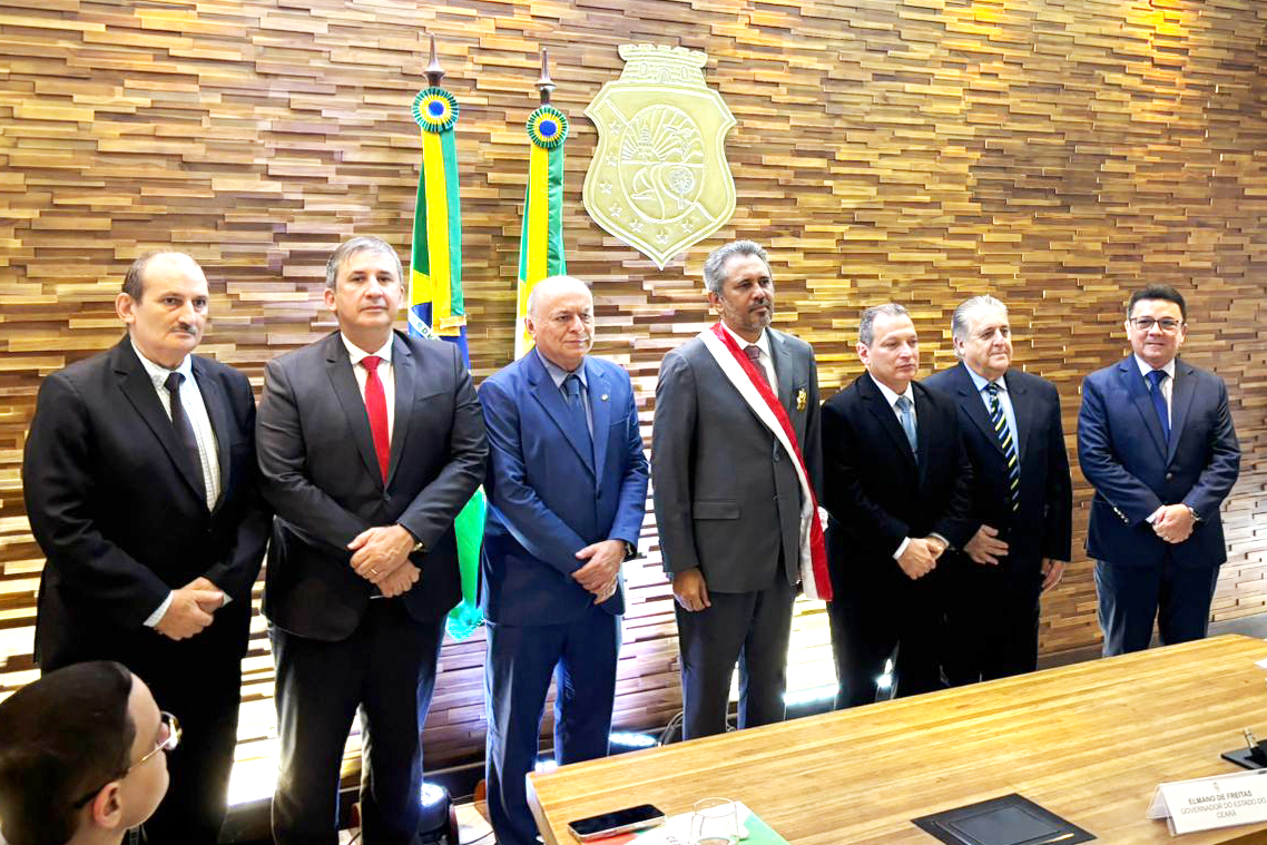  Sete homens de paletó e gravata em pé na frente do brasão do Ceará, o do meio exibe uma faixa e uma medalha