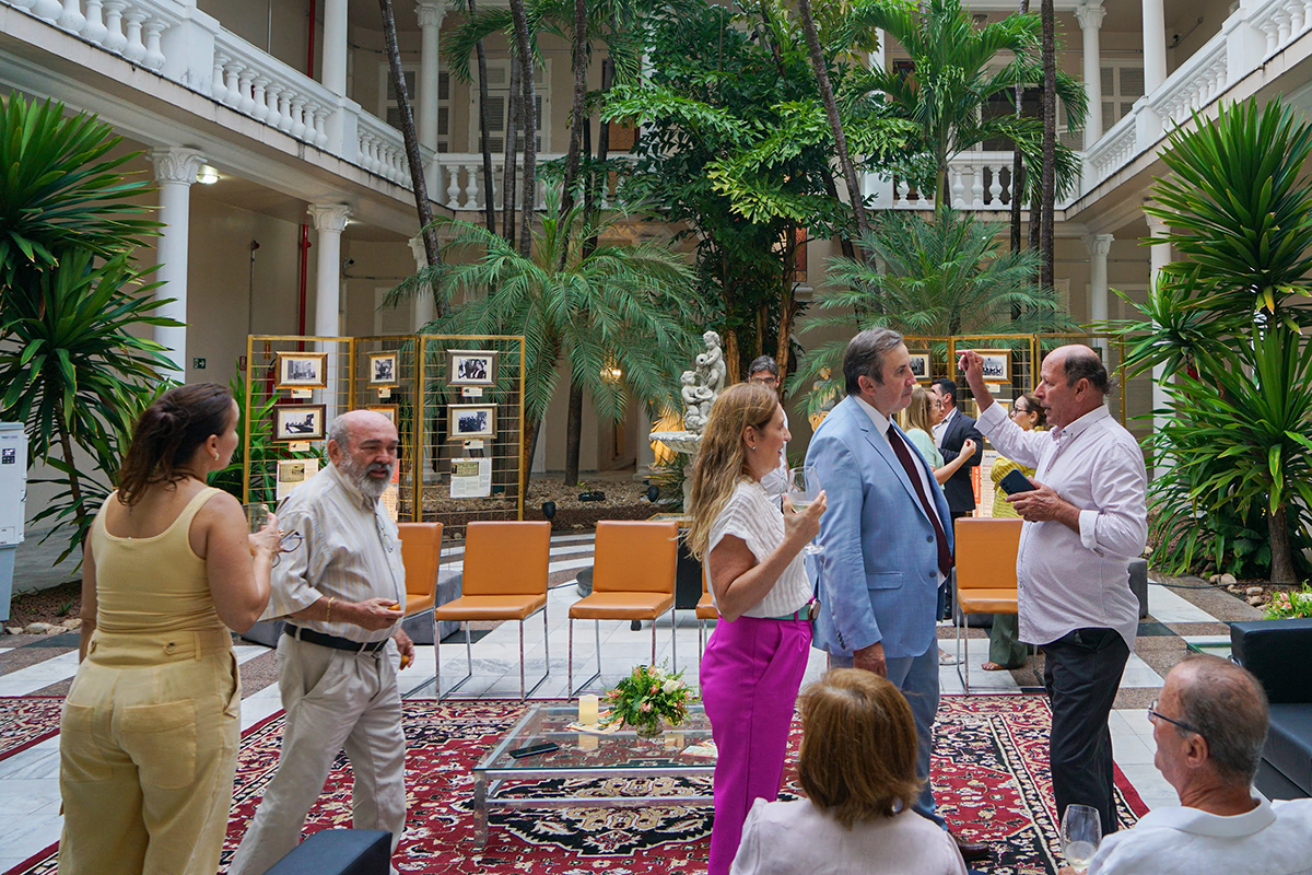 Várias pessoas conversando, algumas em pé e outras sentadas, num jardim interno de uma grande casa, com plantas e palmeiras ao fundo. No centro, há fotografias penduradas em expositores e cadeiras. No chão, um tapete vermelho.