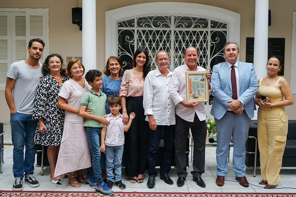 Dez adultos e duas crianças em pé posam pra foto no átrio de um casarão. Um deles exibe um quadro com uma homenagem ao juiz Ubirajara