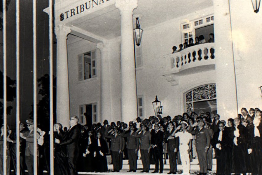 Diversas autoridades em trajes militares, em pé na frente de uma casarão com enormes colunas clássicas, enquanto hasteiam a bandeira