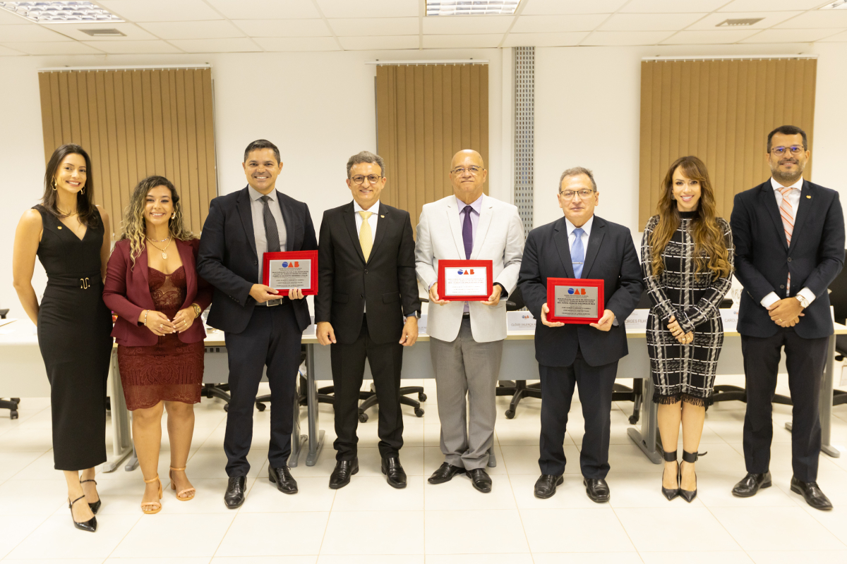 três mulheres e cinco homens em pé numa sala, vestidos com trajes de passeio completo. Três dos homens exibem placas metálicas de homenagem