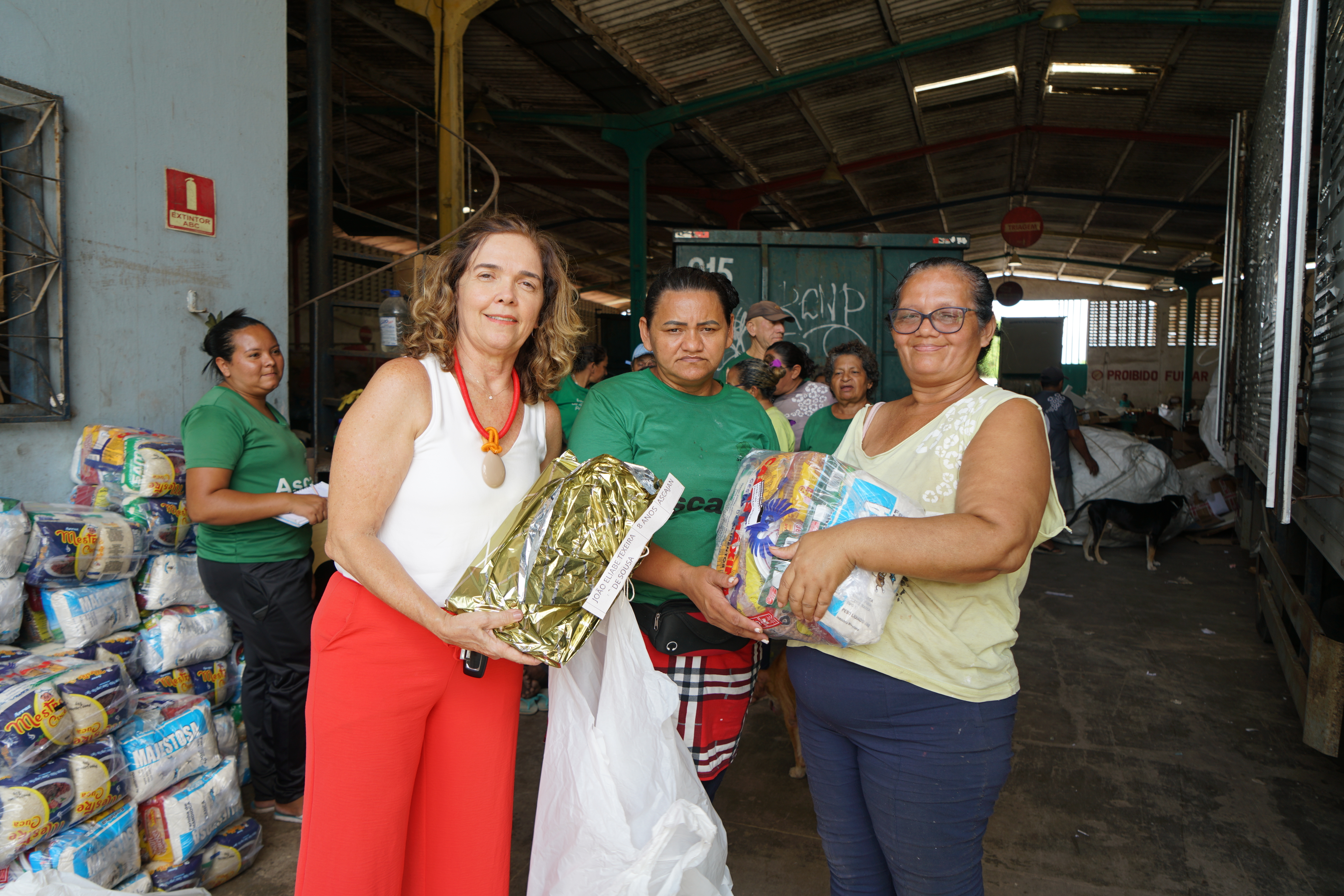Foto de Silvana e duas catadoras. Elas estão segurando presentes e cestas básicas dentro do galpão dos catadores no Jangurussu