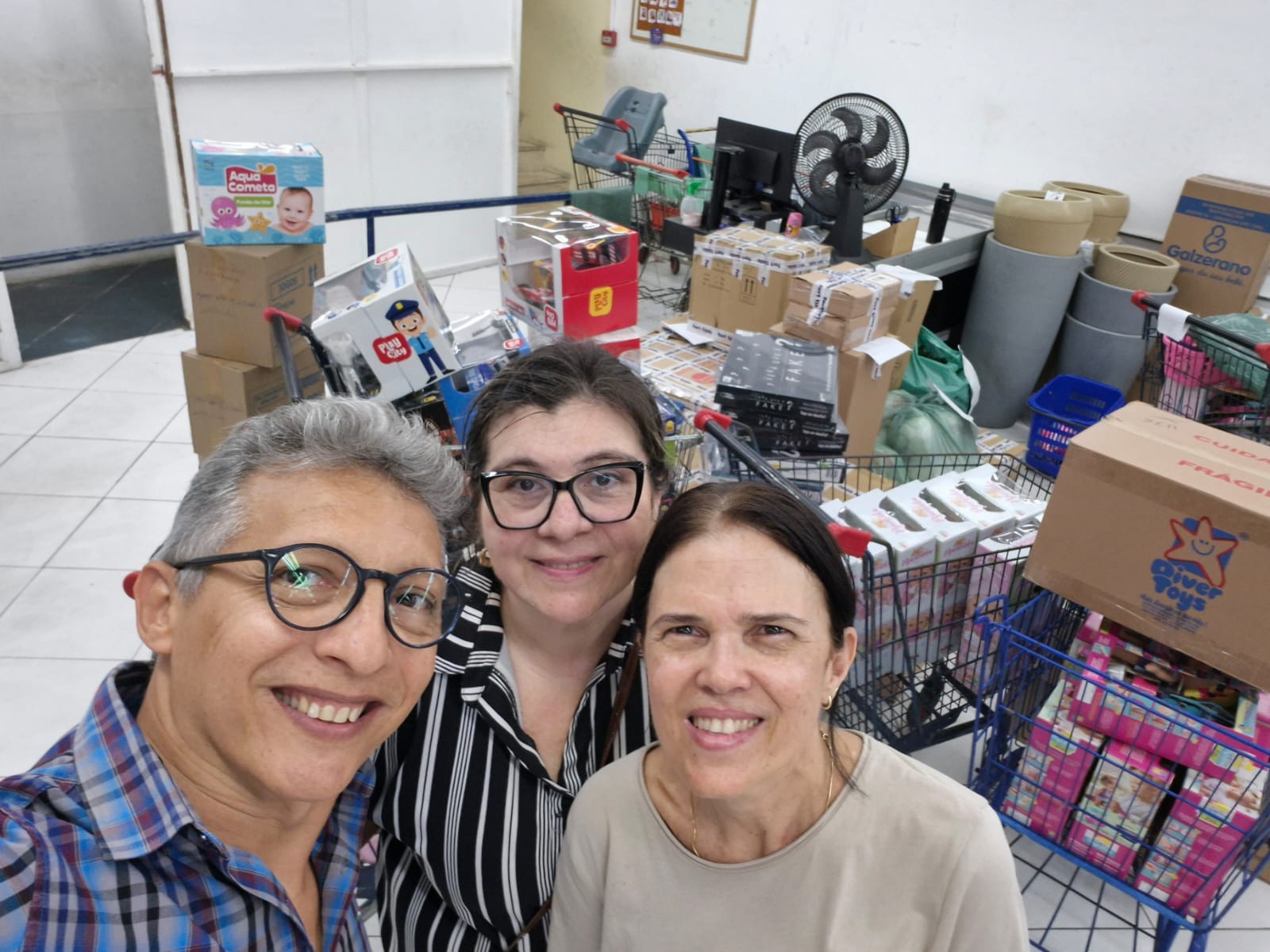 Helano Oliveira, Flávea Andréa Façanha e Leyara Mendonça na compra dos presentes. Eles estão olhando para a foto sorridentes e Helano fazendo uma selfie. Atrás deles tem vários carrinhos com brinquedos.