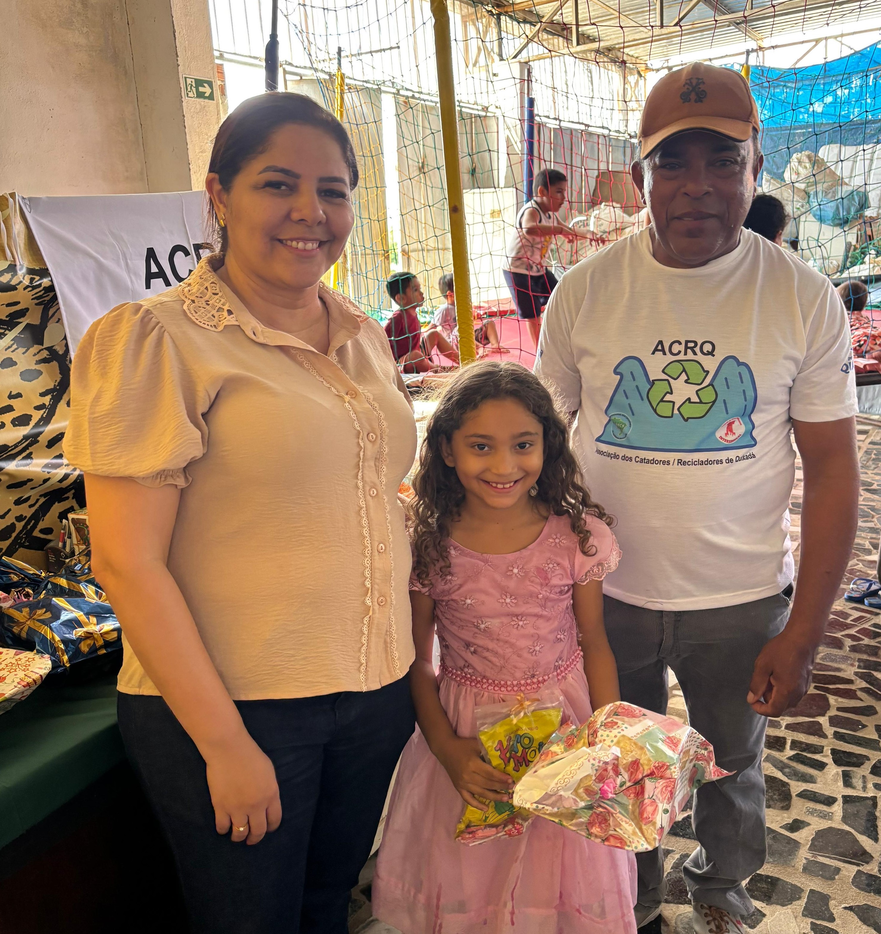 Geyza Nantua realizando a entrega de brinquedos na ACRQ. Ela está entregando uma boneca a uma criança que está ao lado de um parente. Atrás deles tem crianças brincando num pula-pula