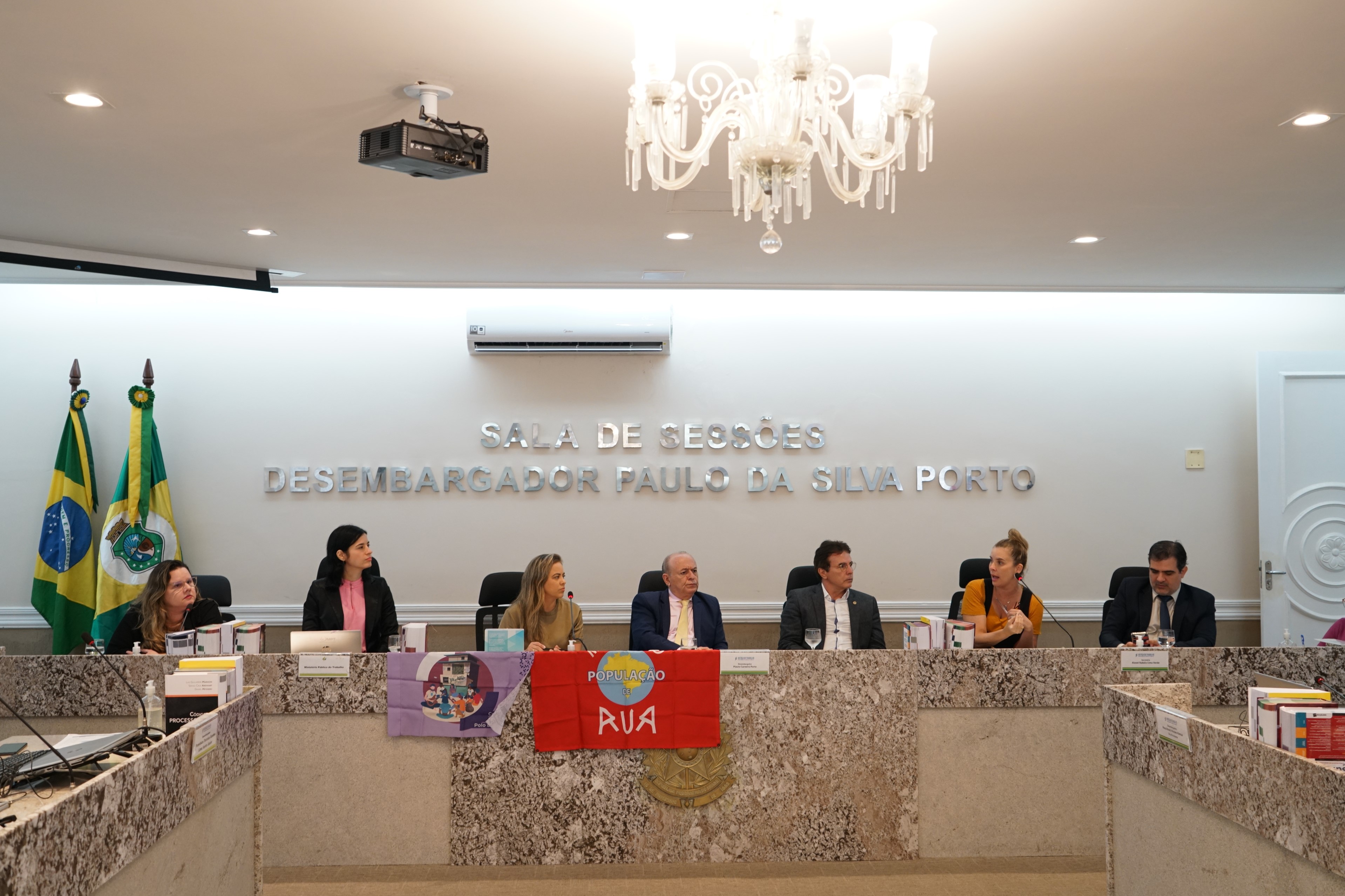Foto de alguns participantes da reunião que estão sentados nas cadeiras dos desembargadores do Pleno. Nesse momento, a defensora Mariana Lobo está falando no microfone e os outros participantes estão escutando