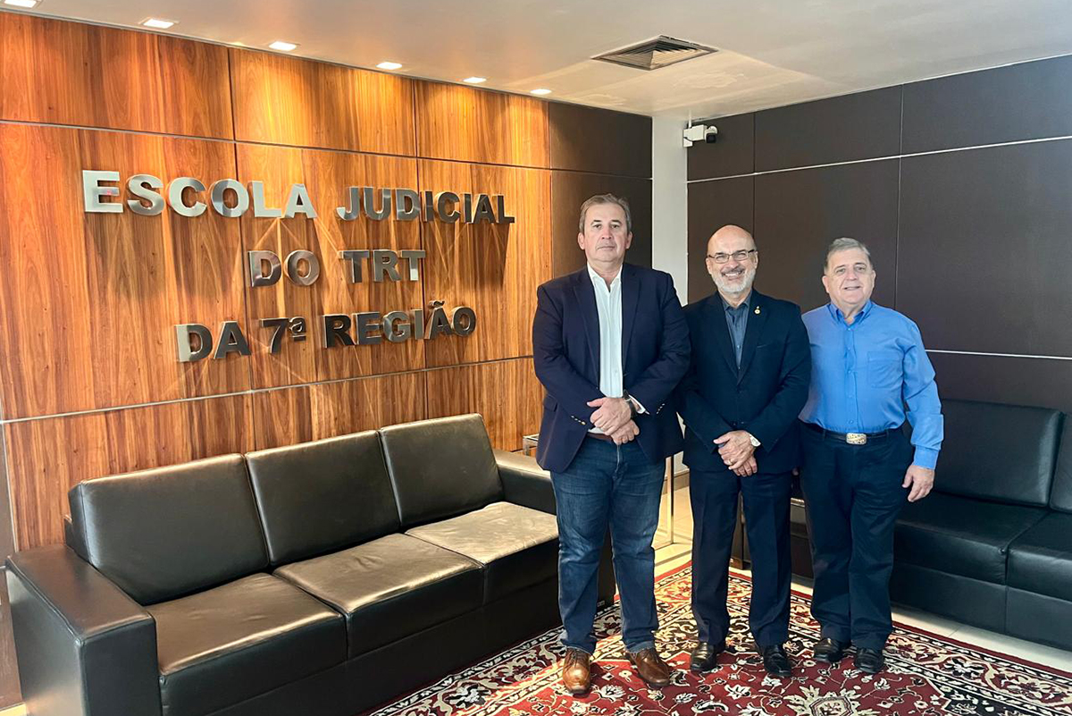 3 homens de blazers azuis em pé posam pra foto na frente do letreiro da Escola Judicial, entre a parede e um sofá preto