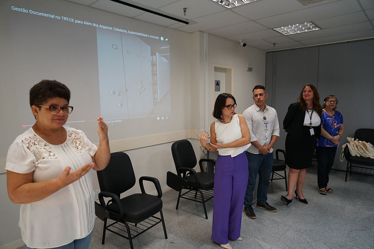 mulher em pé fala apontando pra um telão na parende, que exibe um vídeo. Ao lado dela, uma intérprete de Libras faz sinais com as mãos