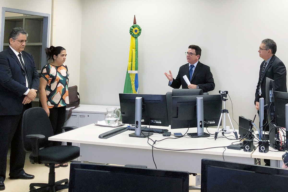 Corregedor ao centro da sala de audiências gesticulando para dois juízes e uma servidora; todos estão em pé