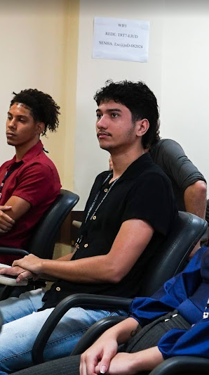 Foto do estagiário Caio Ronei sentado na sala de treinamento assistindo à exposição. Ele veste blusa preta e calça jeans.