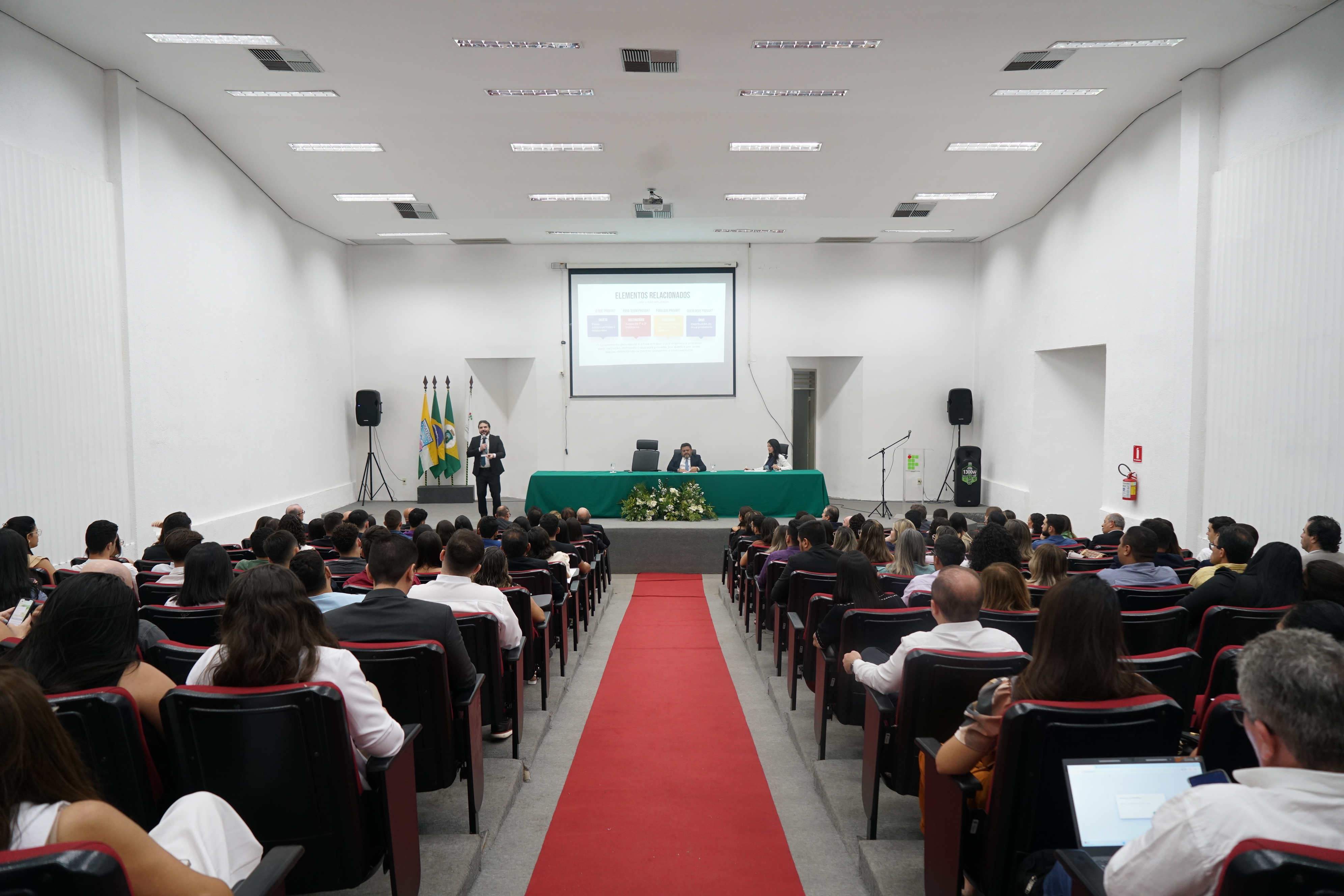 Foto do auditório. Há uma passadeira vermelha, as cadeiras estão cheias e a mesa está com dois participantes