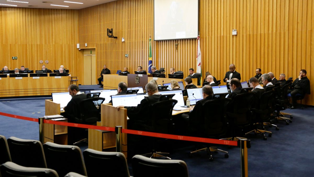 Foto do plenário do TST, onde constam vários magistrados e magistradas de toga preta. Eles estão sentados, em frente a notebooks e estão posicionados num semi-círculo. O fundo do ambiente é amadeirado. 