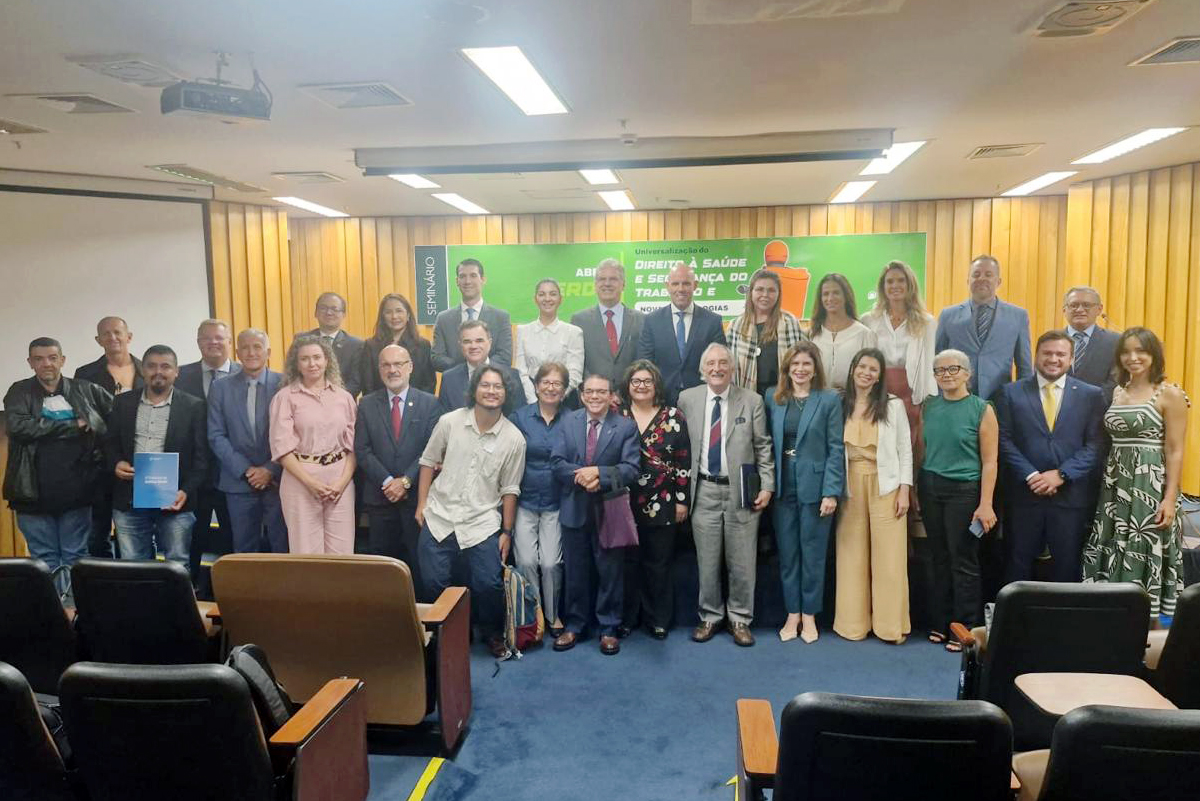  dezenas de pessoas posam em pé num auditório em frente à uma faixa verde com o título do Seminário