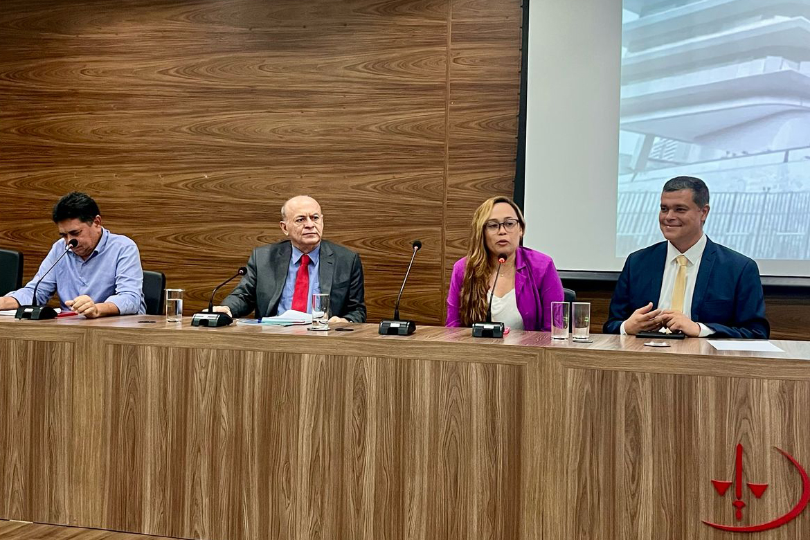 Uma mulher fala ao microfone ao lado de três homens sentados na mesa e abertura do palco com o prédio do MPT no telão ao fundo