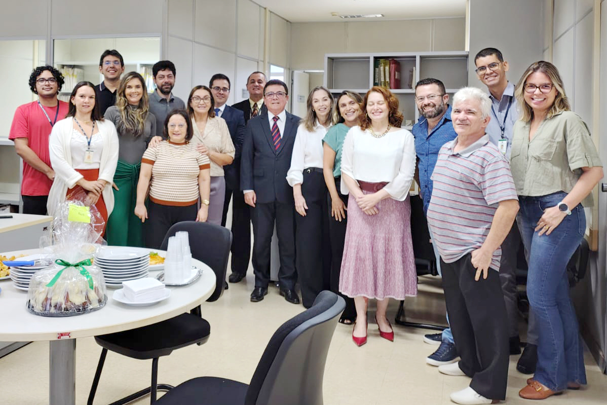  Várias pessoas em pé posam pra foto na secretaria da vara, com estantes de processos ao fundo e uma mesa com comidas à frente