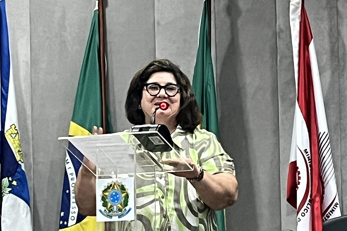 mulher de vestido verde fala numa tribuna com bandeiras ao fundo