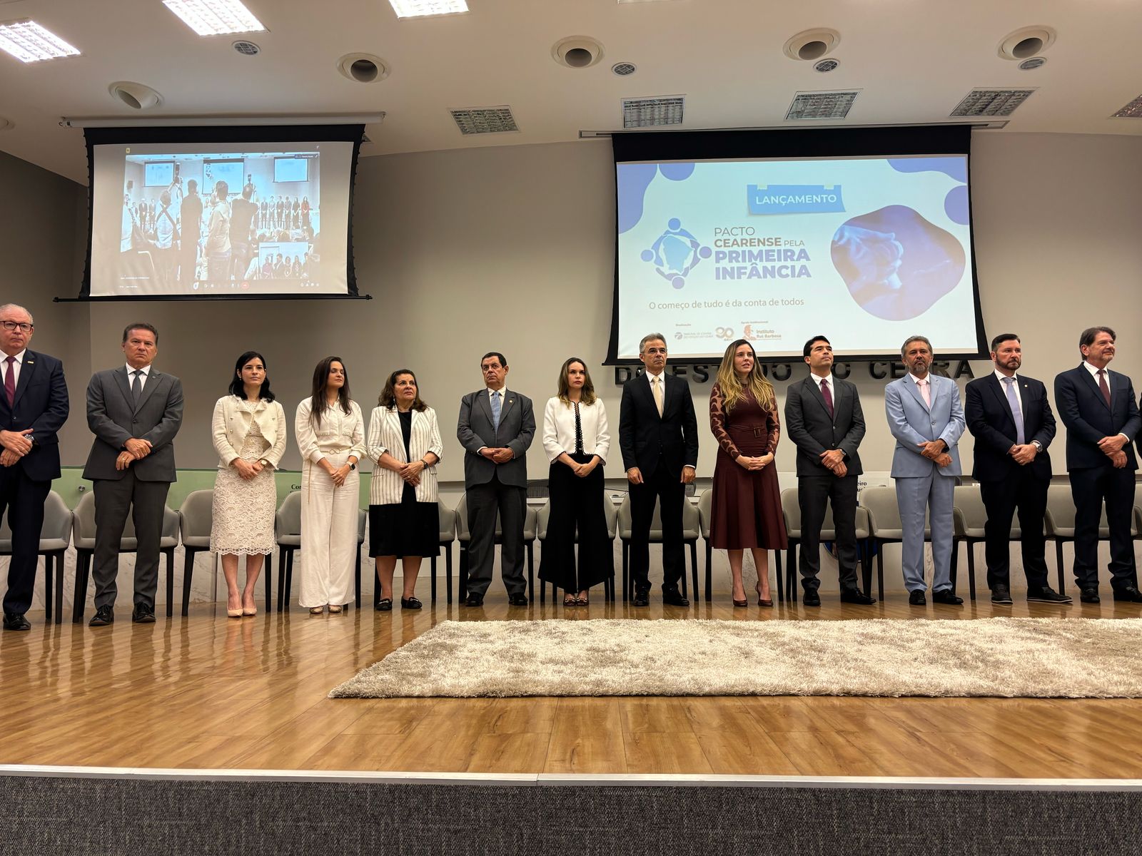 autoridades em pé no palco do evento com logomarca do evento ao fundo no telão