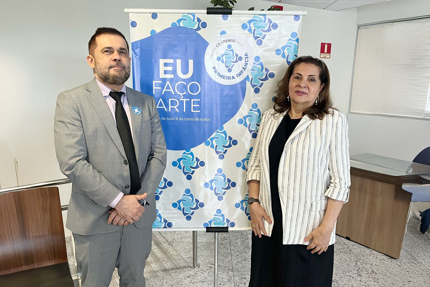 homem de terno e mulher de blazer posam para foto ao lado do banner com o logotipo do evento na cor azul