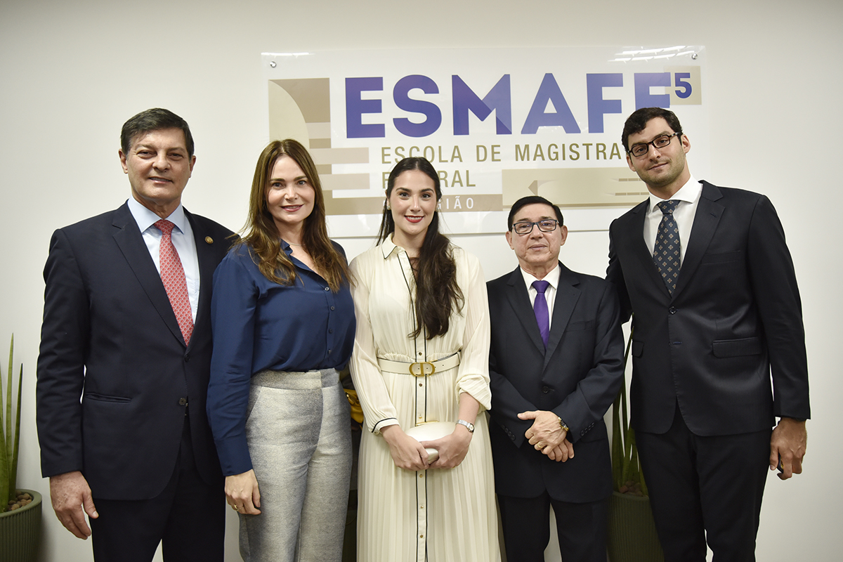 dois homens de terno e gravata e duas mulheres de vestido numa sala com parede branca e um grande emblema pendurado com o nome ESMAFE em azul
