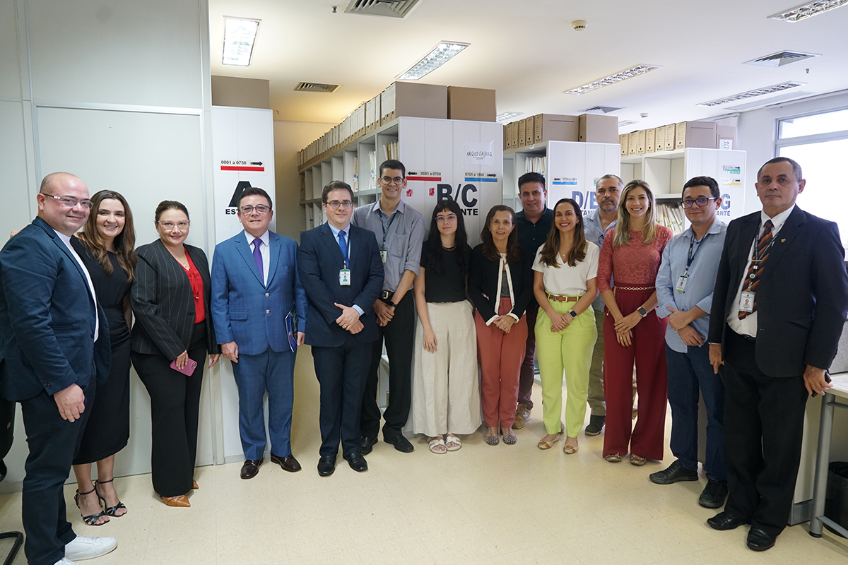 Um grupo de catorze pessoas está posando para uma foto em uma secretaria de vara. Há oito homens e seis mulheres. Alguns dos homens vestem ternos, enquanto outros usam camisas casuais. As mulheres vestem roupas variadas, incluindo blusas listradas e calças jeans. Ao fundo, há armários de escritório e uma porta.