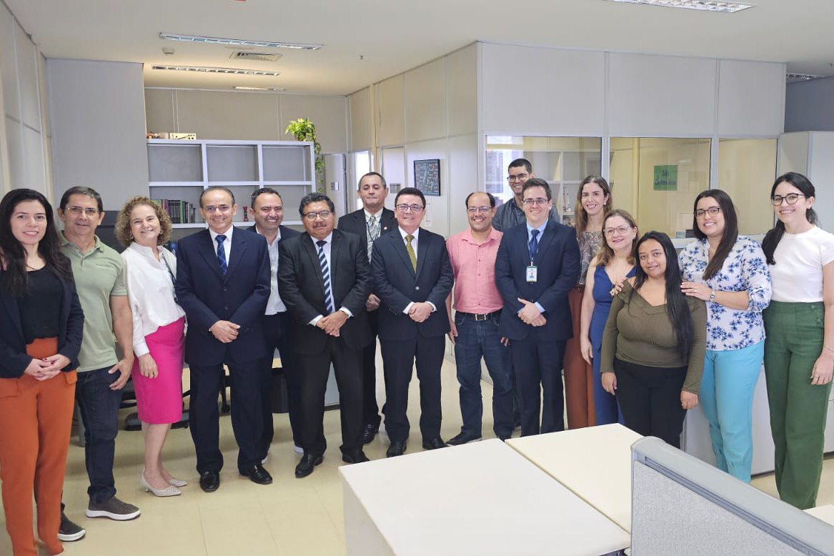 Um grupo maior, composto por dezesseis pessoas, posa para uma fotografia em um ambiente de escritório. Há dez homens e seis mulheres no grupo. Os homens vestem uma variedade de trajes, incluindo ternos em tons de azul e cinza com camisas e gravatas, camisas sociais de manga comprida em cores como verde claro e rosa, e calças jeans. As mulheres vestem diferentes tipos de roupas, como blusas e calças em tons de laranja, verde e azul com estampas florais, vestidos em tons de azul e saias em cores como rosa. Uma das mulheres veste óculos de grau. O fundo da imagem mostra um ambiente de escritório com divisórias claras, algumas plantas em vasos e mesas de trabalho com tampos claros e divisórias cinzas. A iluminação é artificial, vinda do teto. As pessoas estão dispostas em duas fileiras, com algumas na frente e outras atrás, todas olhando para a câmera.