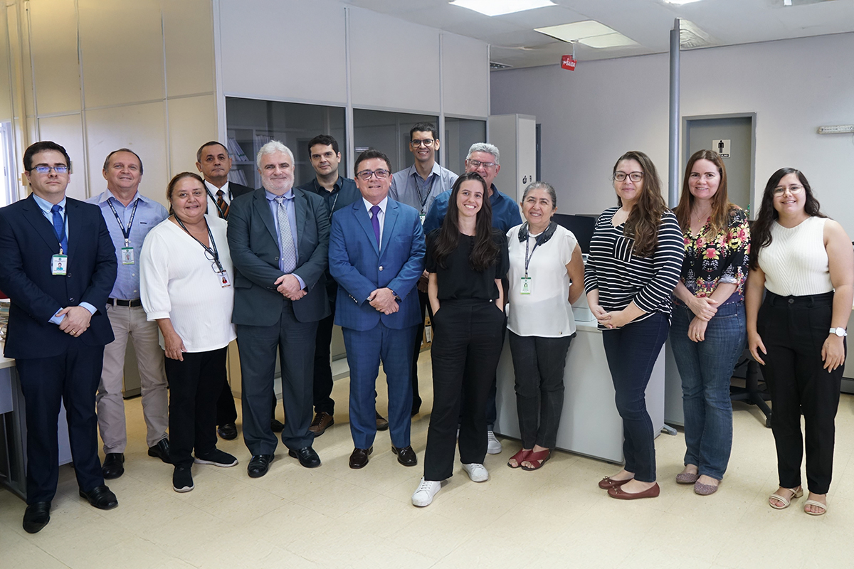 Um grupo de catorze pessoas está posando para uma foto em um ambiente de escritório. Há oito homens e seis mulheres, todos olhando para a câmera. Alguns dos homens vestem ternos e gravatas, enquanto outros usam camisas sociais. As mulheres vestem uma variedade de roupas, incluindo vestidos e calças. Ao fundo, vê-se prateleiras de arquivos e outros equipamentos de escritório.