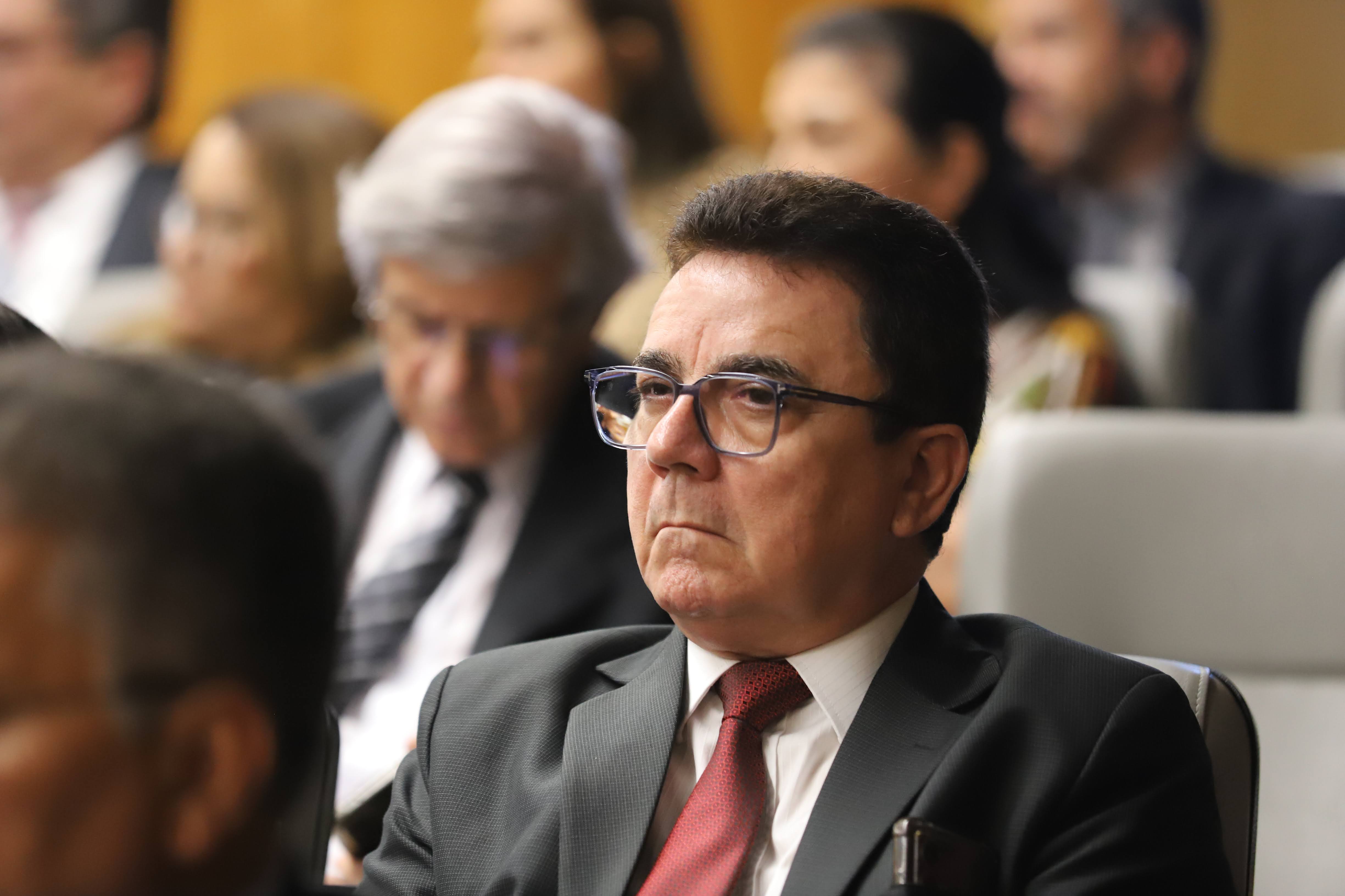  Um homem de meia-idade (des. João Carlos Uchoa), com cabelos escuros e óculos de armação azul, está sentado em uma cadeira, prestando atenção. Ele veste um terno escuro com uma camisa branca e uma gravata vermelha estampada. O ambiente ao fundo sugere que ele está em um evento formal, possivelmente uma palestra ou reunião.