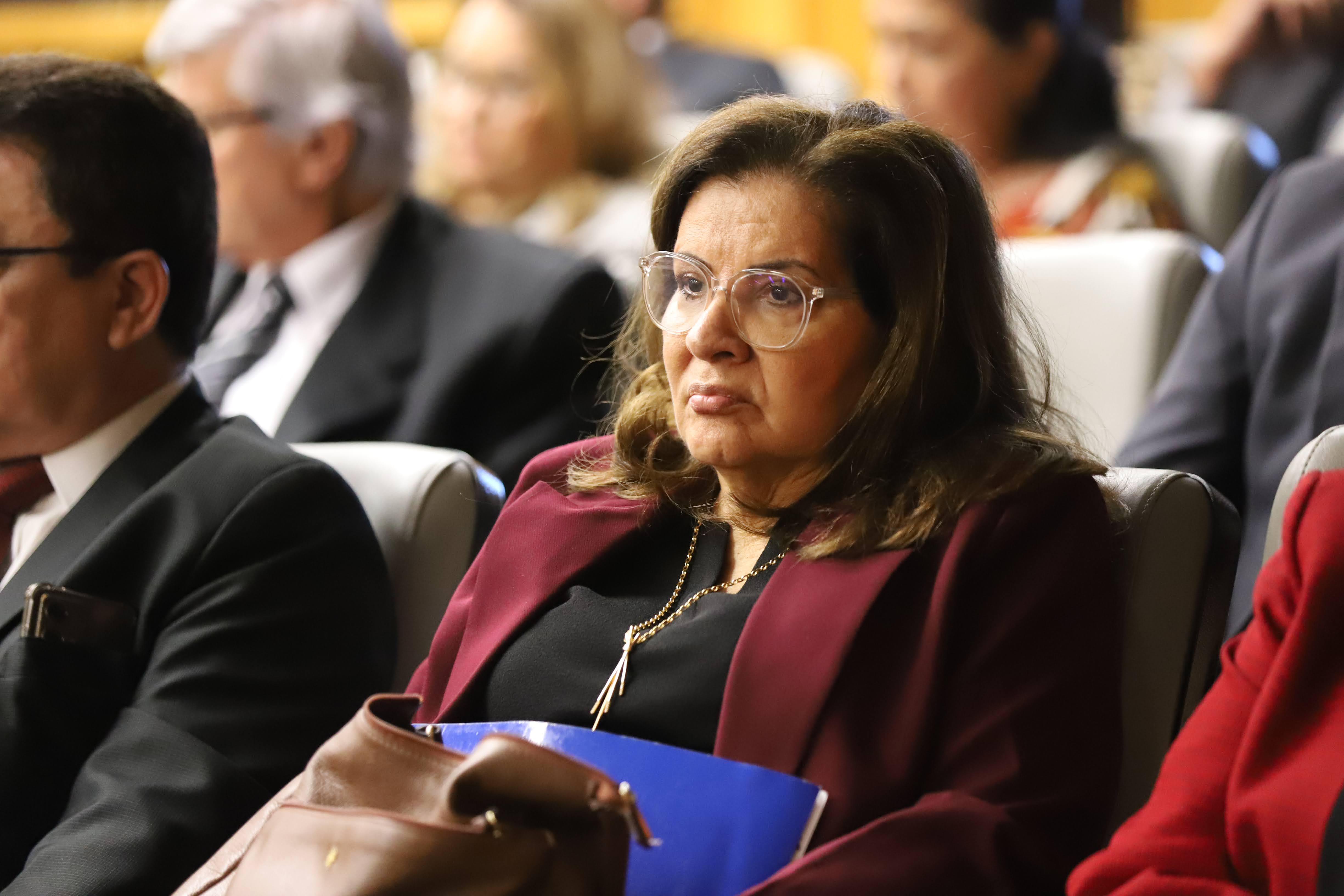  Uma mulher de meia-idade (Desa. Fernanda Uchoa), com cabelos castanhos escuros e óculos claros, senta-se atenta em uma sala formal. Ela veste um blazer vinho sobre uma blusa preta, adornada com um colar dourado. Ao seu lado, uma bolsa marrom e uma pasta azul repousam em seu colo.