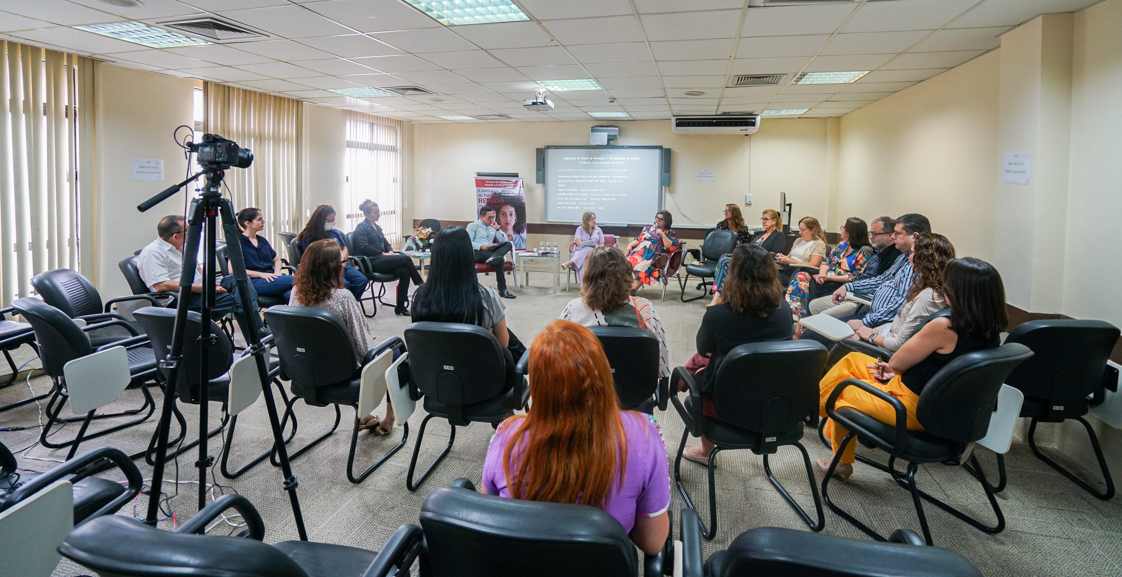 Uma sala com várias pessoas sentadas em cadeiras pretas, voltadas para frente. Na frente, há uma mesa com algumas pessoas sentadas e falando, além de um projetor e uma tela com texto. Uma câmera de vídeo em um tripé está à esquerda.