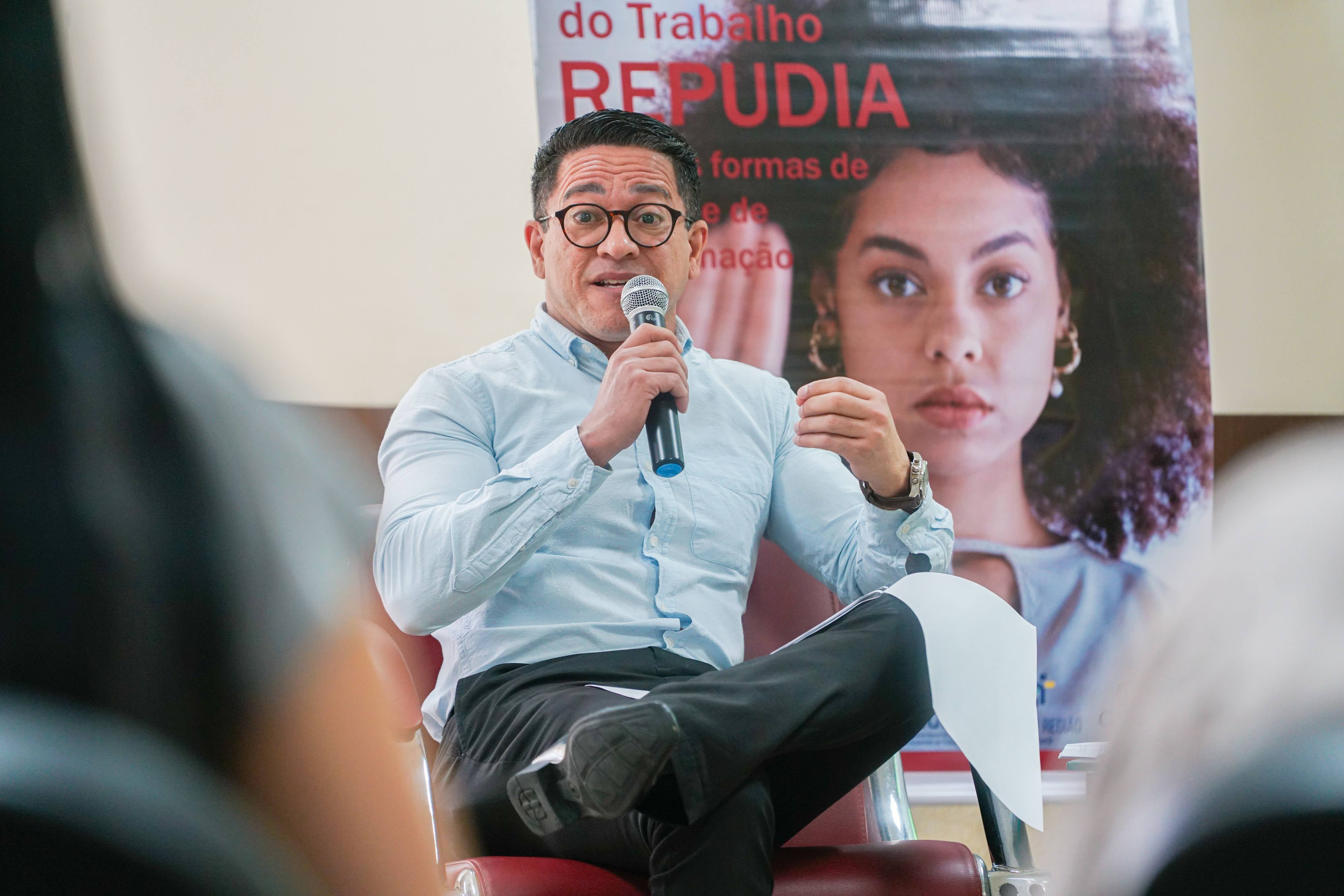 Um homem de pele clara, com óculos e camisa azul clara, está sentado em uma cadeira vermelha, falando em um microfone. Ao fundo, desfocado, vê-se um cartaz com uma mulher de pele morena.