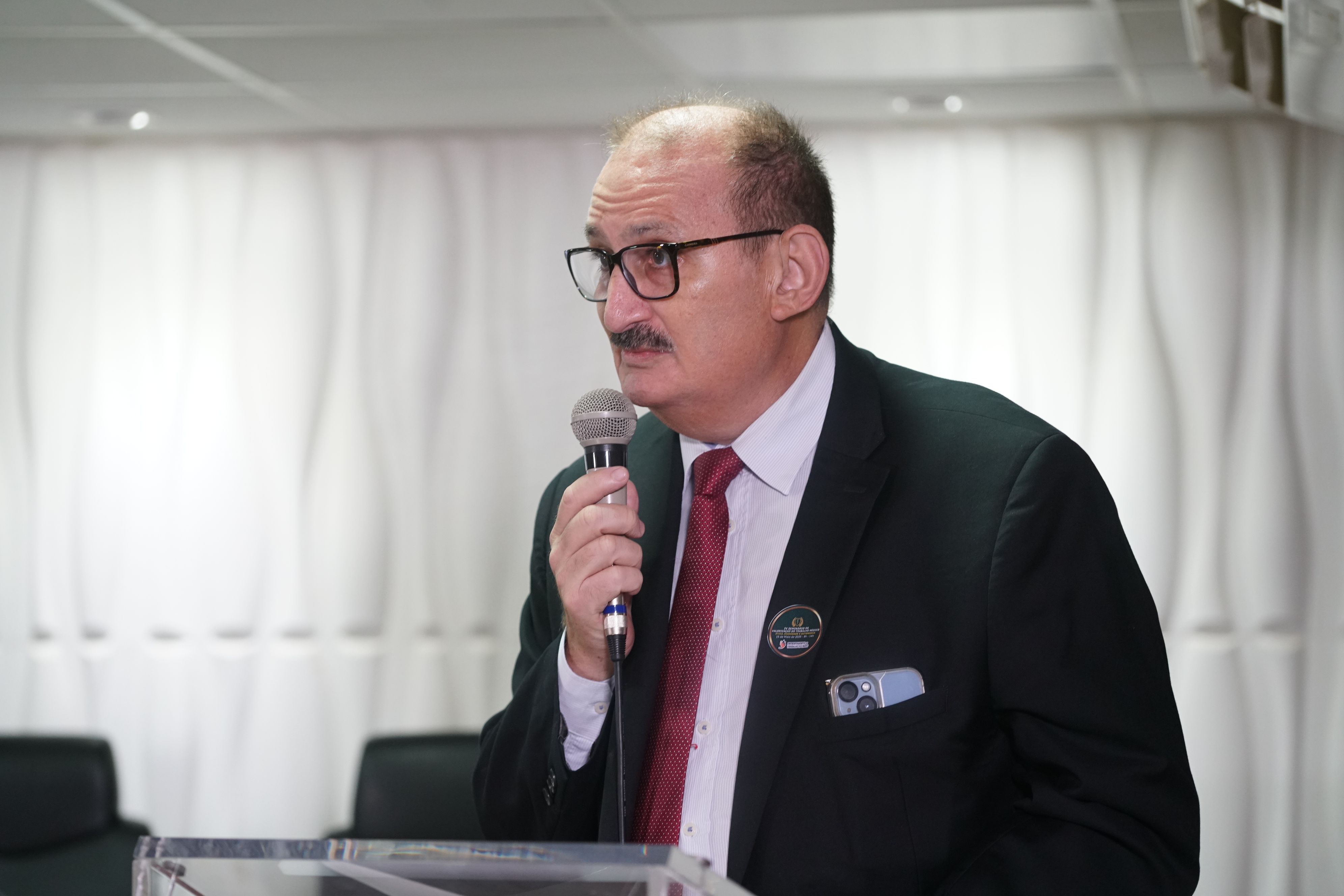 Um homem com pouco cabelo escuro, bigode e óculos fala em um microfone. Ele veste um terno escuro, uma camisa branca e uma gravata vermelha. Ele está em pé atrás de uma tribuna transparente.