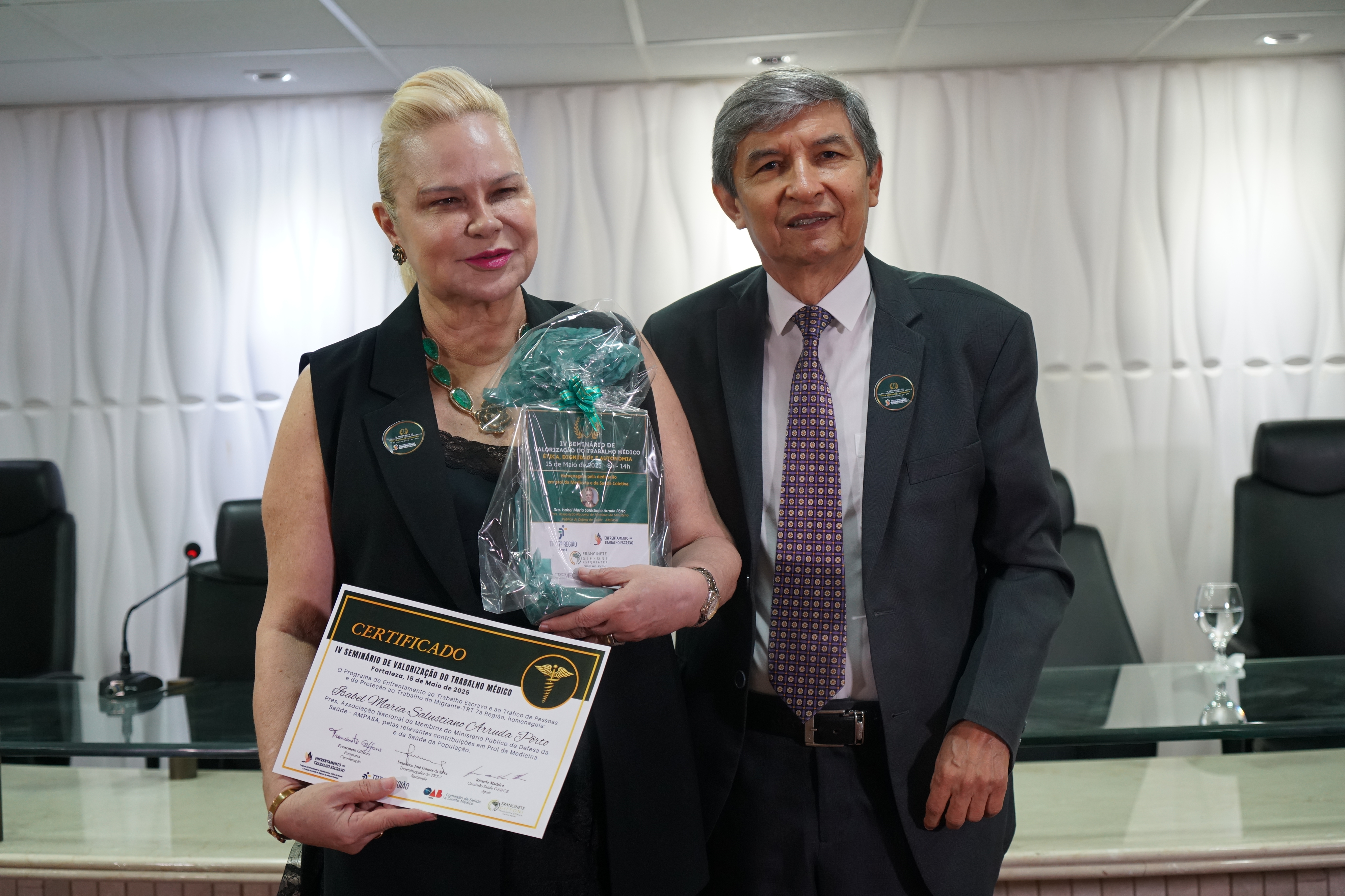 Uma mulher de pele clara com cabelos loiros presos segura um certificado e um presente embrulhado. Ela está ao lado de um homem com cabelos grisalhos e escuros que veste um terno escuro e uma gravata estampada. Eles estão em uma sala com uma mesa e cadeiras ao fundo. O certificado tem texto em português e apresenta um símbolo de caduceu.