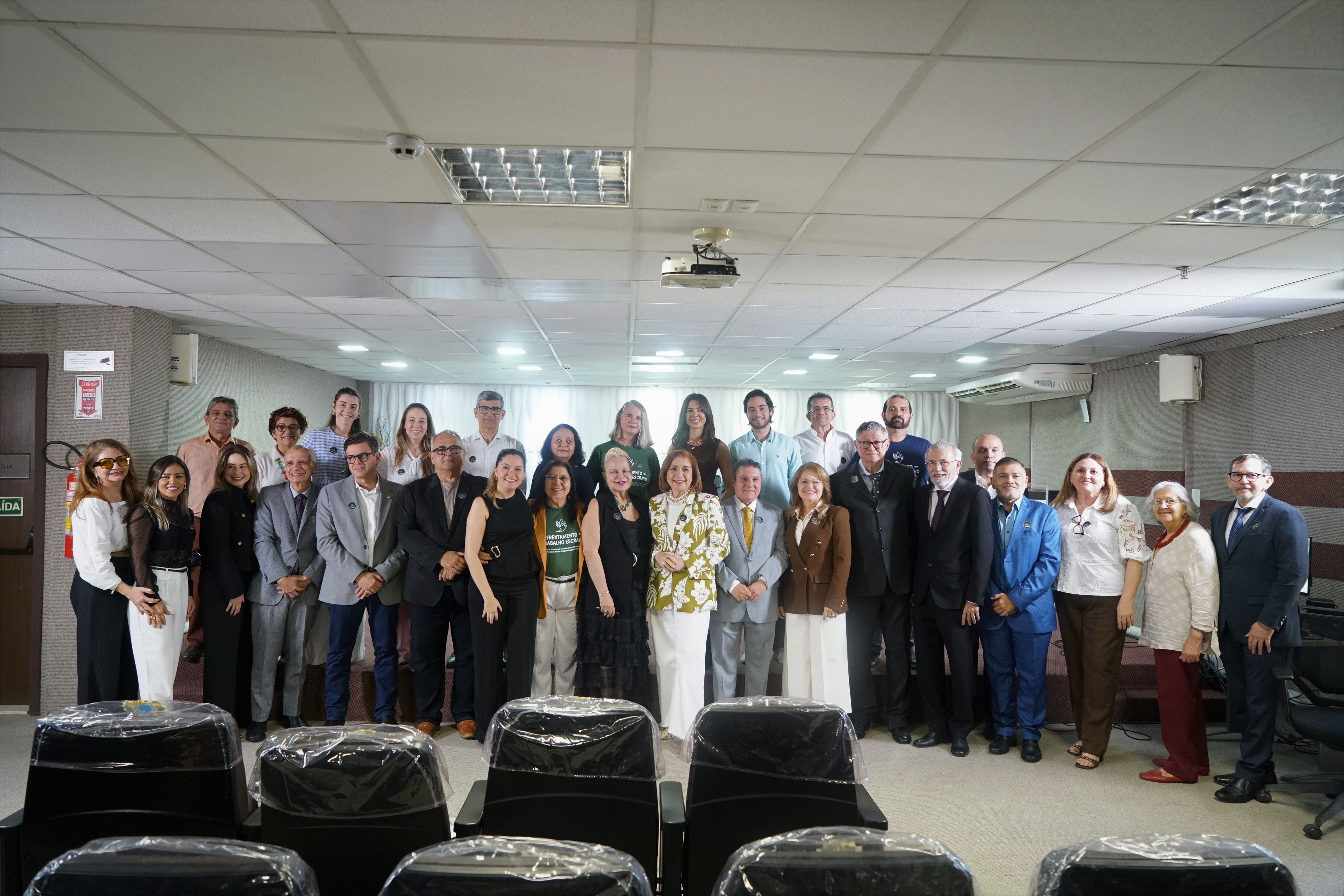 Um grupo grande de aproximadamente trinta pessoas de várias idades e aparências está em pé, posando para uma foto em uma sala com teto rebaixado e iluminação fluorescente. A maioria veste trajes casuais de negócios. Há uma porta com uma placa "SAÍDA" à esquerda e cadeiras cobertas em primeiro plano.