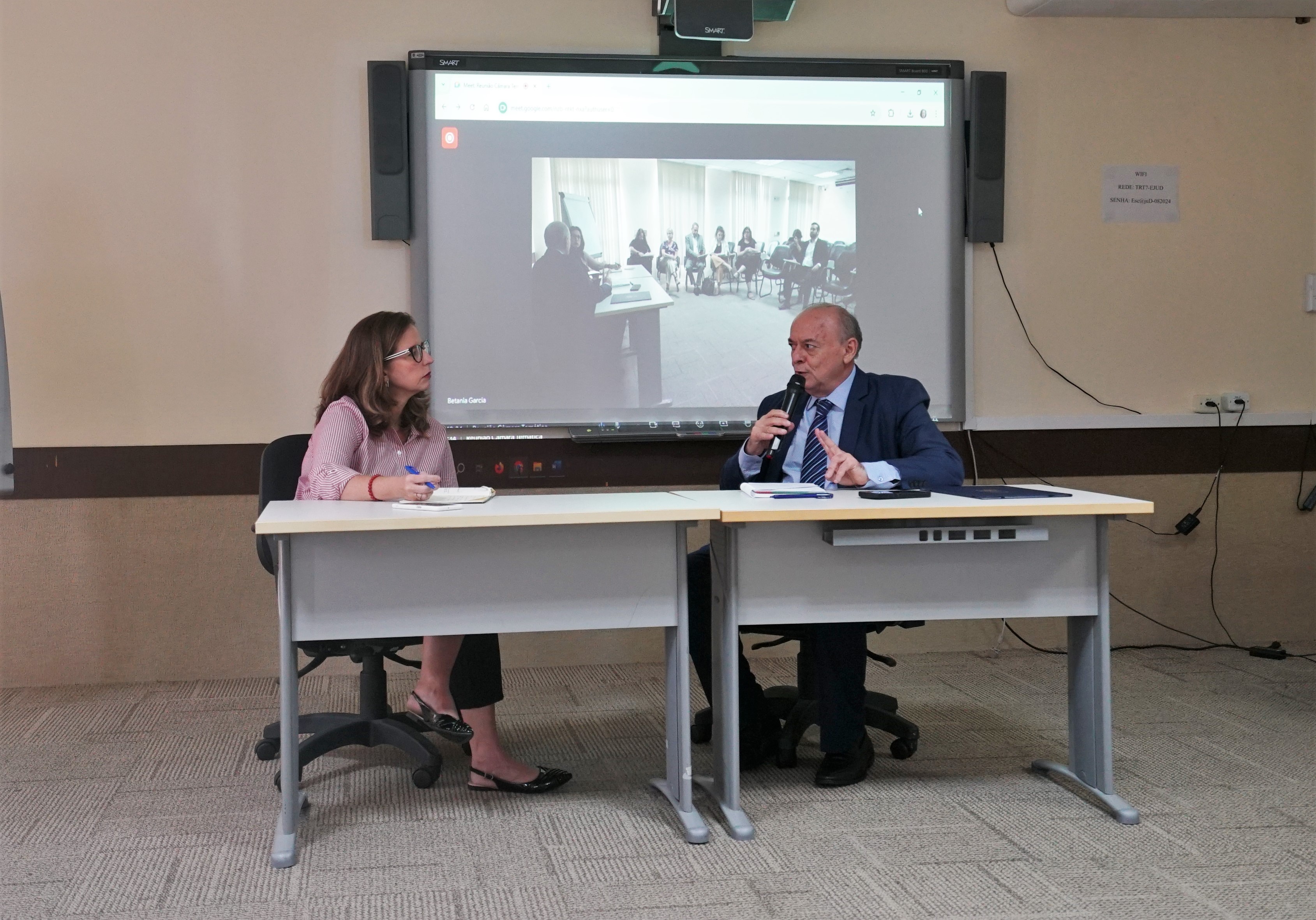 Foto do Dr. Parente e uma participante. Ambos estão sentados em duas mesas. No fundo, há um telão reproduzindo a imagem do restante dos participantes