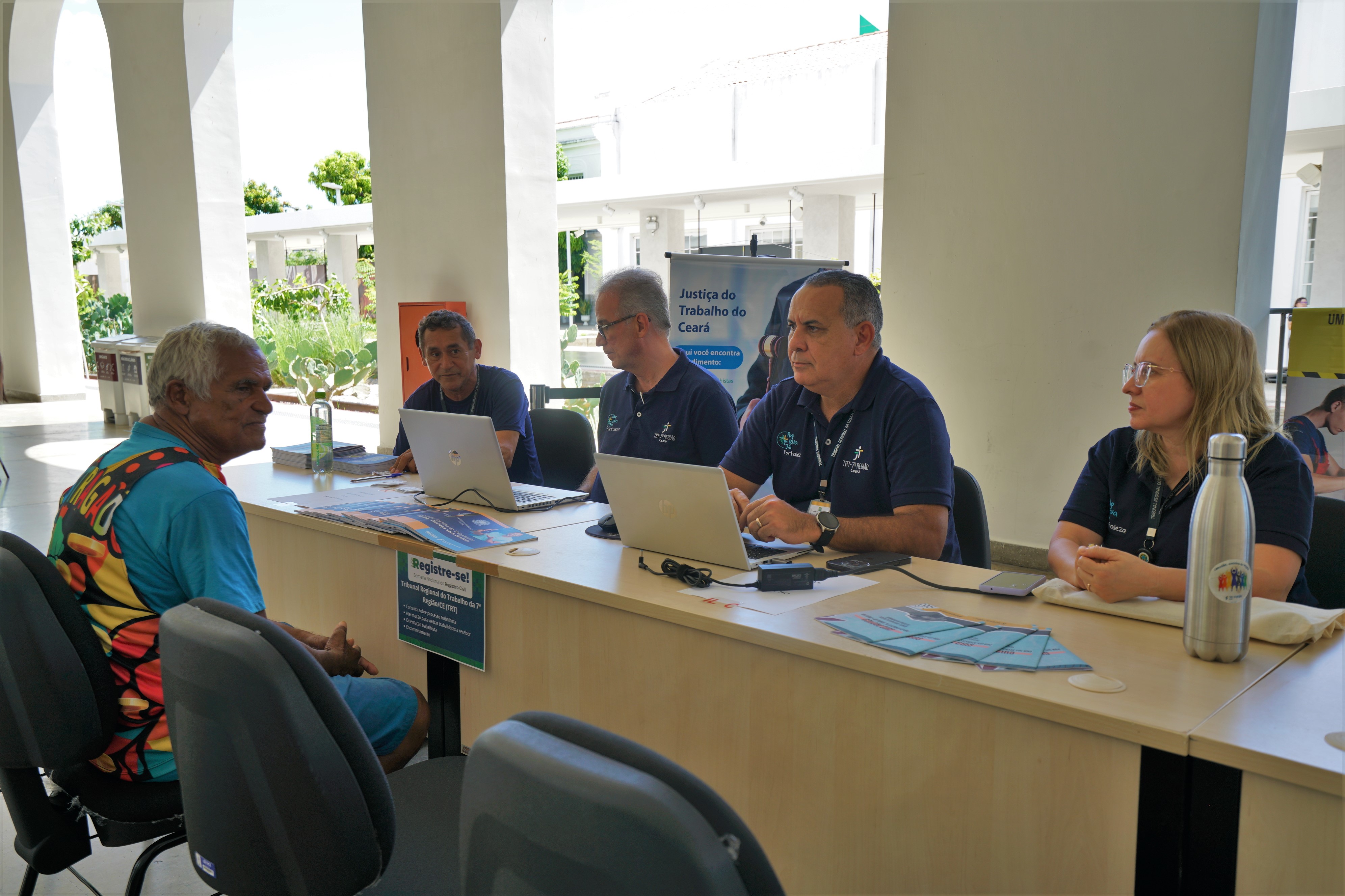 A imagem mostra cinco pessoas interagindo em uma mesa sob uma estrutura aberta com colunas brancas. À esquerda, um homem de pele mais escura com cabelos grisalhos está sentado, voltado para a mesa. Quatro pessoas de pele clara estão sentadas atrás da mesa, cada uma com um laptop e materiais diversos. Um banner com a inscrição "Justiça do Trabalho do Ceará" é visível na mesa, sugerindo um evento ou serviço relacionado ao trabalho.