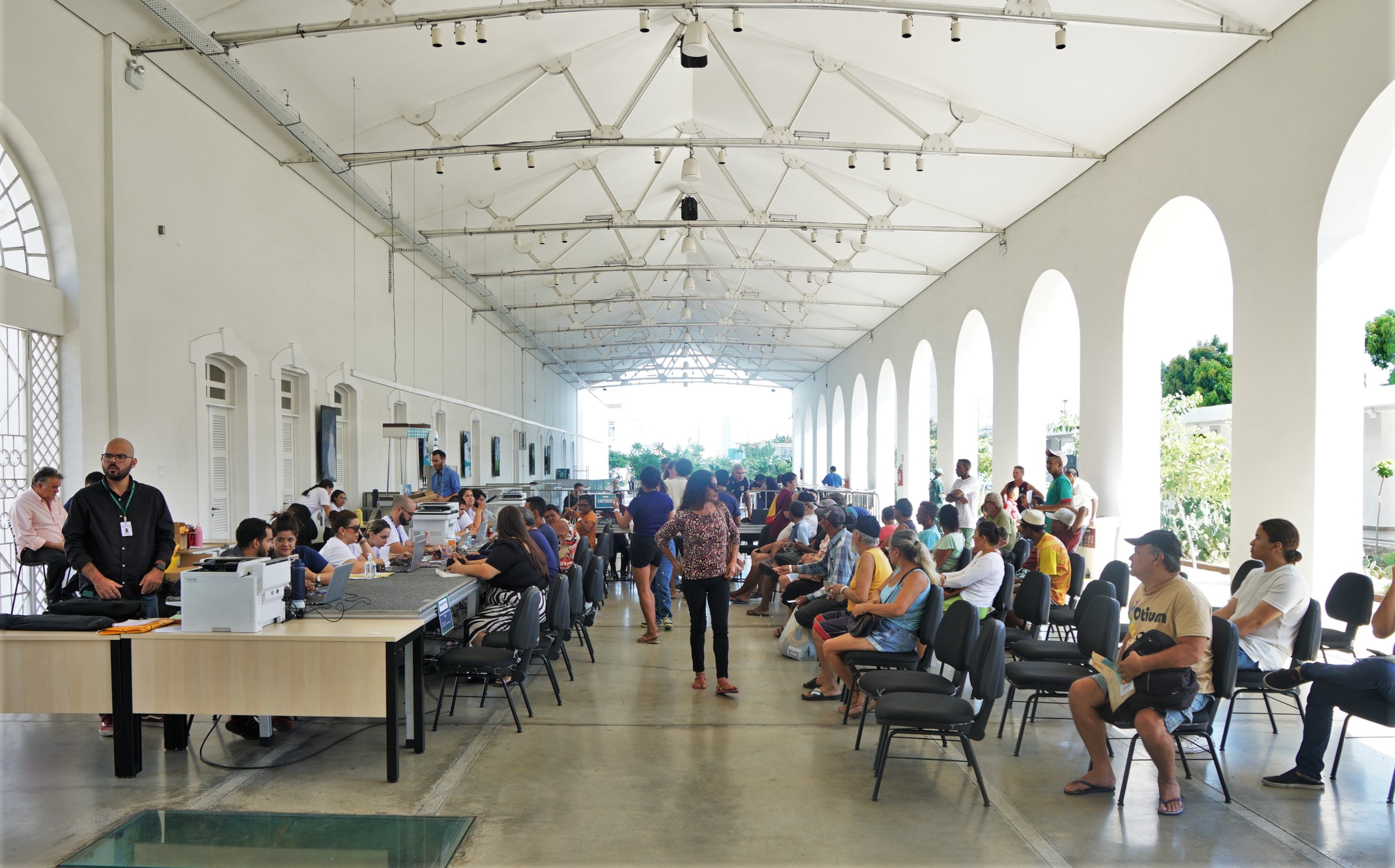 A imagem panorâmica mostra um grande espaço interno com teto alto de estrutura metálica branca e paredes brancas com arcos. Diversas pessoas estão espalhadas pelo local, algumas sentadas em cadeiras pretas voltadas para mesas, enquanto outras estão em pé ou caminhando. A iluminação natural entra pelas aberturas nas laterais, criando um ambiente claro e movimentado.