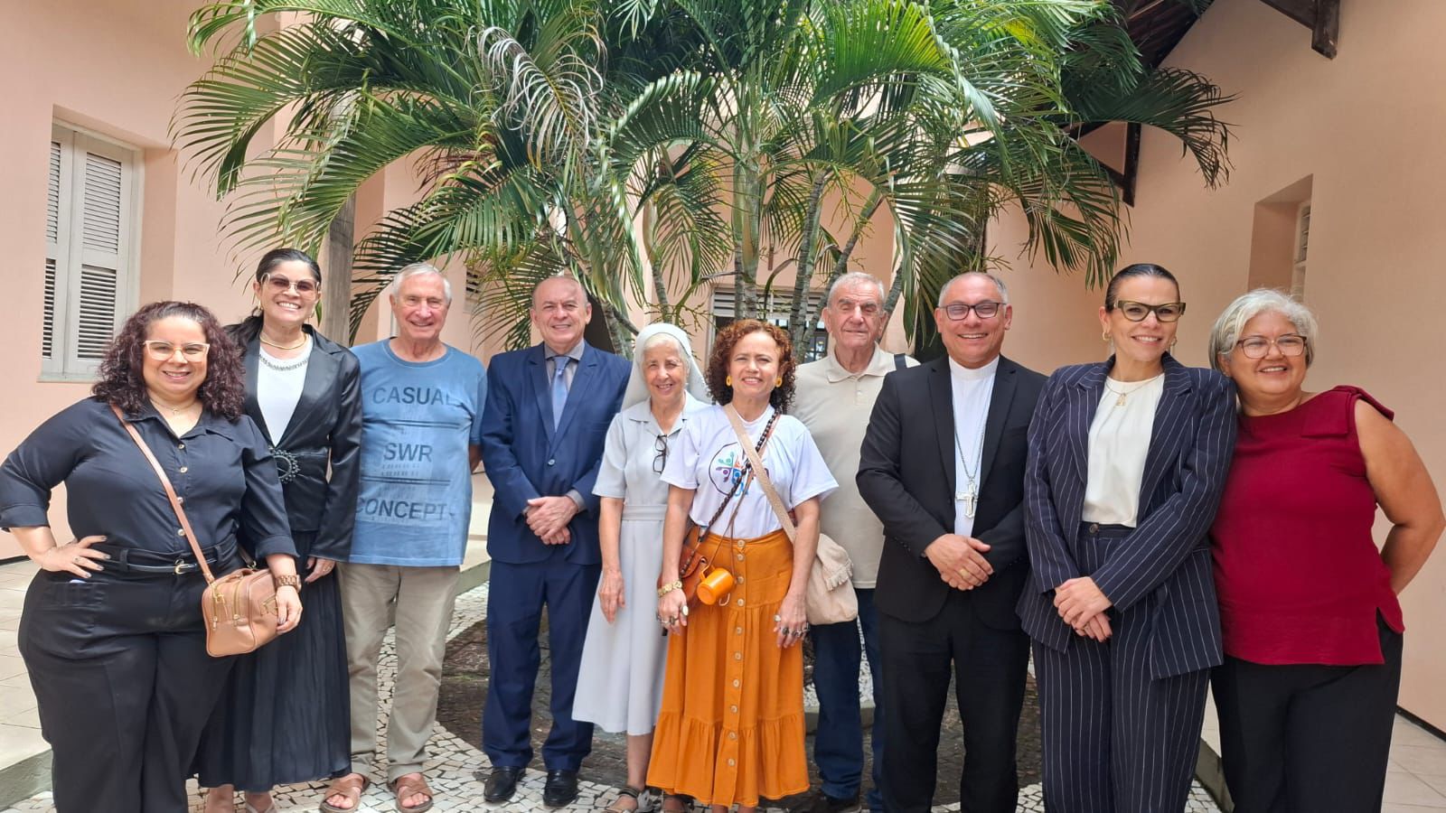  A fotografia mostra um grupo diversificado de dez pessoas, incluindo homens e mulheres de várias idades, posando juntos ao ar livre. Ao centro da imagem, há uma grande planta com folhas de palmeira, e o fundo é composto por uma parede de cor clara. O grupo está sorrindo e olhando para a câmera.
