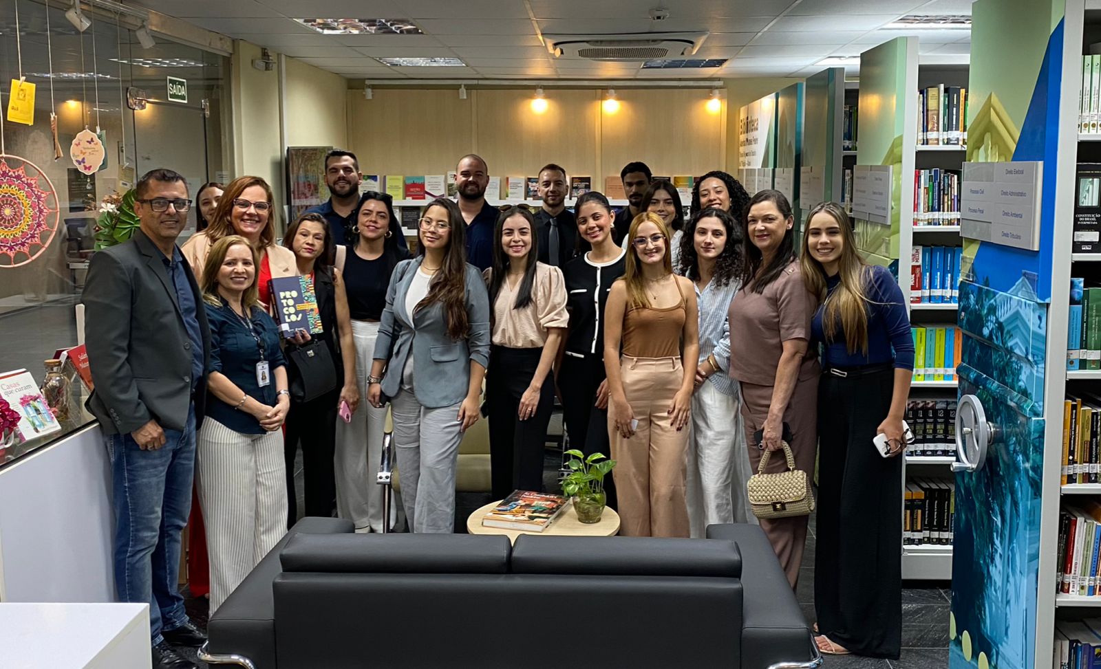 Uma imagem mostra um grupo diverso de aproximadamente 20 pessoas em uma biblioteca, a maioria mulheres e algumas com bolsas, dispostas informalmente. A sala é iluminada e organizada com estantes de livros ao fundo, sofás e mesas baixas, além de algumas decorações e vasos de plantas. Todos estão olhando para a câmera e sorrindo.