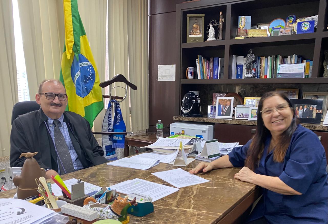 A imagem mostra duas pessoas sentadas em uma mesa de escritório com uma superfície de mármore marrom. À esquerda, o desembargador Francisco José Gomes um homem de pele clara com bigode, óculos e vestindo um terno escuro sobre uma camisa clara com gravata estampada, olha para a câmera. Atrás dele, à esquerda, está uma bandeira do Brasil parcialmente visível, e à direita, uma estante de madeira escura repleta de livros, objetos decorativos e fotografias emolduradas. À direita da mesa, a doutora Francinete Giffoni, médica psiquiatra, uma mulher de pele clara, com óculos e vestindo uma blusa azul, sorri para a câmera. A mesa está coberta com papéis, canetas, um calendário e outros objetos de escritório. O ambiente sugere um escritório formal.