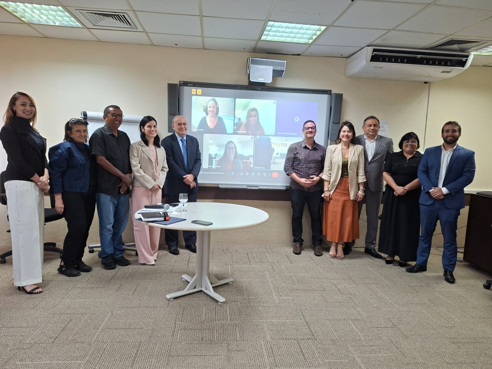 Um grupo de dez pessoas está reunido em uma sala com uma tela interativa mostrando quatro participantes em uma videoconferência. Seis pessoas estão em pé, lado a lado, voltadas para a câmera, enquanto quatro outras estão visíveis na tela ao fundo. Há uma mesa redonda branca com objetos e duas cadeiras vazias à esquerda do grupo. O ambiente tem iluminação artificial, paredes claras e um carpete estampado.