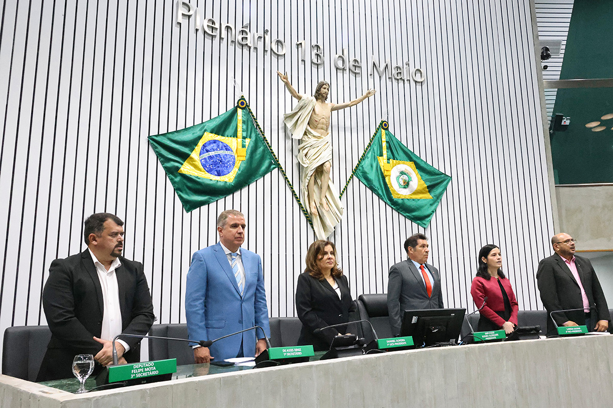 A imagem mostra o plenário da Assembleia Legislativa do Ceará, conhecido como "Plenário 13 de Maio". Vários indivíduos, aparentemente em uma sessão ou cerimônia oficial, estão posicionados atrás de uma bancada com microfones e placas de identificação. Ao fundo, na parede, destaca-se a inscrição "Plenário 13 de Maio" em letras douradas, acompanhada de uma escultura de Jesus Cristo crucificado e duas bandeiras (do Brasil e do Ceará) posicionadas simetricamente. A sala possui um design moderno com painéis verticais claros na parede.