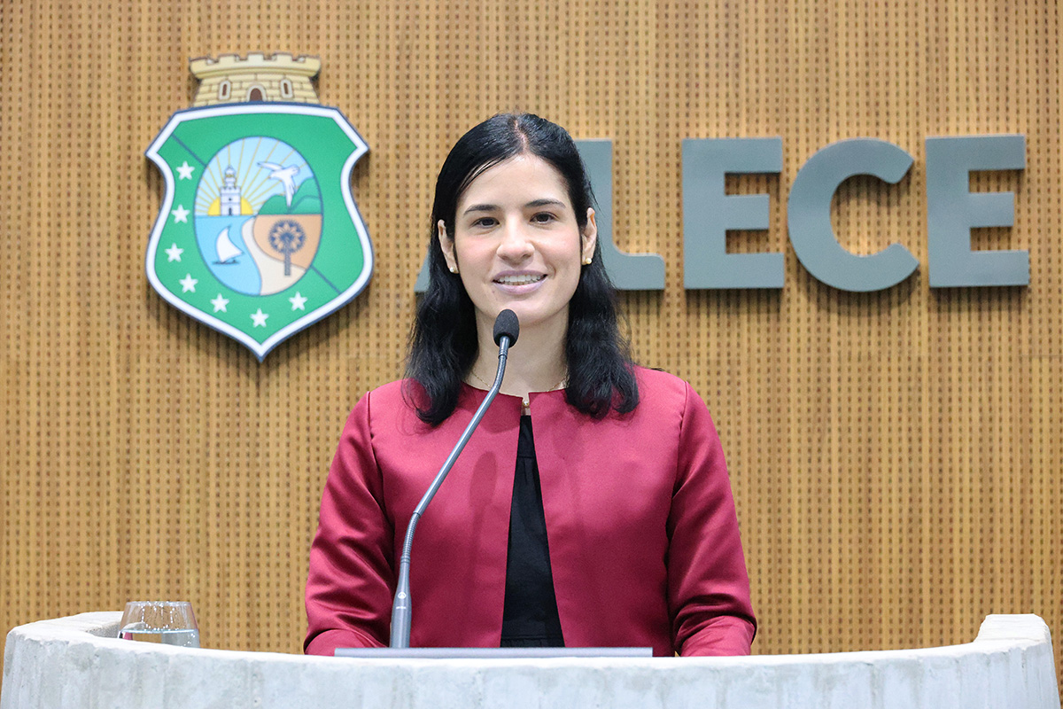 A imagem apresenta uma mulher de pele clara e cabelos escuros, vestindo um blazer vermelho sobre uma blusa preta, sorrindo enquanto está posicionada atrás de um púlpito claro com um microfone. Ao fundo, na parede revestida de madeira, destaca-se um brasão colorido com a inscrição "ALECE" em letras grandes à direita. A mulher parece estar em um ambiente oficial, possivelmente discursando ou participando de um evento formal. Um objeto de vidro, talvez um copo, é visível à esquerda do púlpito.