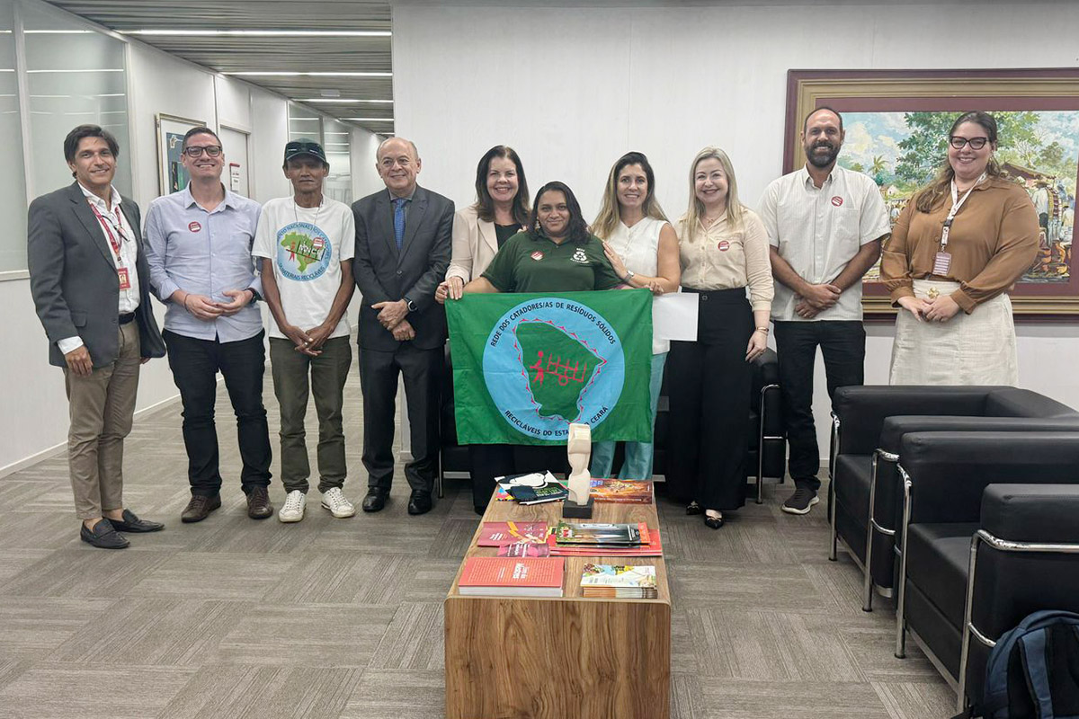 Um grupo de dez pessoas, aparentemente nove adultos e um jovem, posa sorrindo para uma foto em um ambiente interno com iluminação artificial. Ao centro, duas mulheres seguram uma bandeira verde com um círculo azul e desenhos em tons de verde e vermelho. As pessoas à esquerda da bandeira são quatro homens e uma mulher, enquanto à direita estão um homem e três mulheres. Ao fundo, observa-se uma parede clara com duas molduras, sendo uma com uma pintura paisagística e outra menor com uma imagem indistinta. Na frente do grupo, há uma mesa de madeira clara com objetos coloridos sobre ela, e à direita, parte de um sofá de couro preto é visível. O chão é coberto por um carpete cinza com padrões lineares.