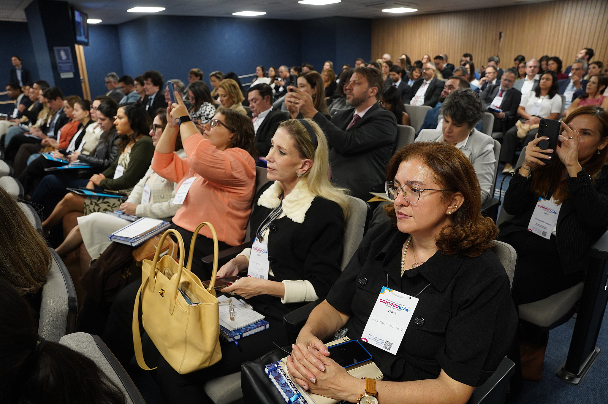 Uma foto média captura uma seção de uma grande plateia sentada em fileiras de cadeiras. Os participantes são diversos em aparência e estão olhando para a frente, alguns segurando telefones para tirar fotos. Muitos estão usando crachás de identificação. O cenário parece ser uma conferência ou apresentação, com paredes azuis escuras ao fundo.