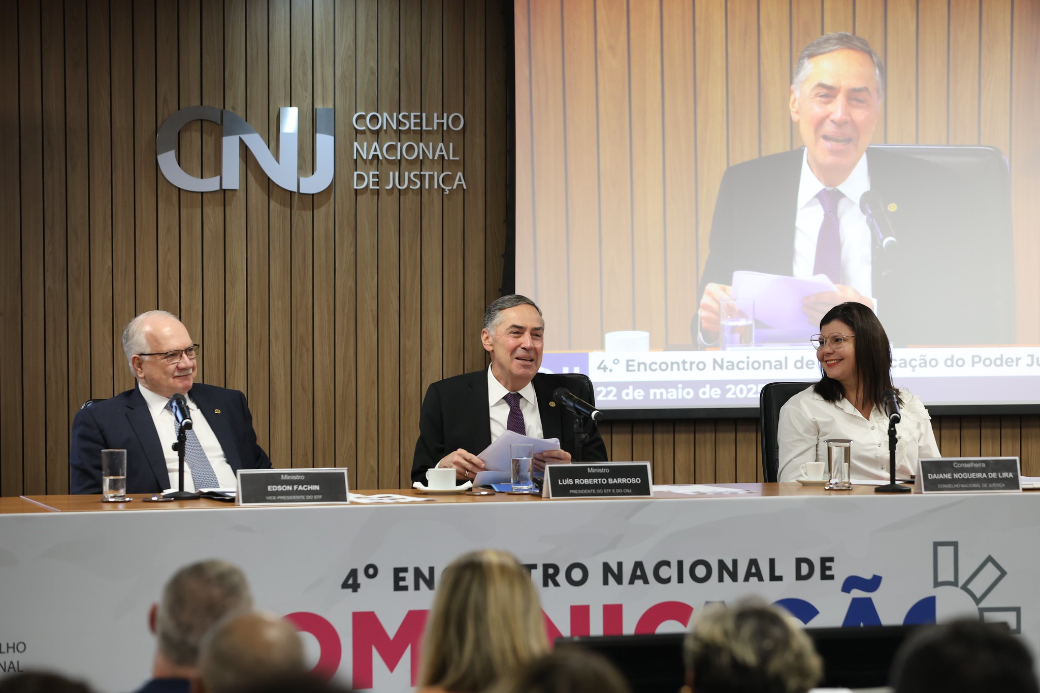 Uma foto ampla mostra um evento formal com um painel de três indivíduos sentados a uma mesa longa em um palco. Atrás deles, há uma parede de madeira com o logotipo "CNJ" e "CONSELHO NACIONAL DE JUSTIÇA" exibido de forma proeminente. Uma tela mostra uma quarta pessoa falando. Os membros do painel têm placas de identificação à frente deles, identificando-os como Edson Fachin, Luis Roberto Barroso e Daiane Nogueira de Lira. A plateia está parcialmente visível no primeiro plano, ouvindo atentamente.