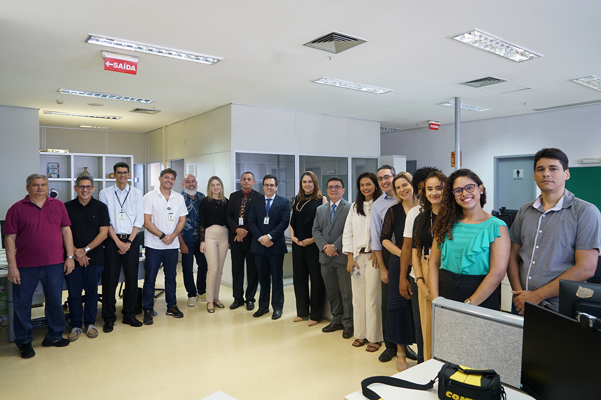 Um grupo de cerca de quinze profissionais, homens e mulheres, está em pé e posa para uma foto em um escritório bem iluminado. Todos estão vestidos com trajes sociais ou executivos, e a maioria sorri para a câmera. Ao fundo, pode-se ver prateleiras e armários de escritório, além de um letreiro vermelho de "SAÍDA".