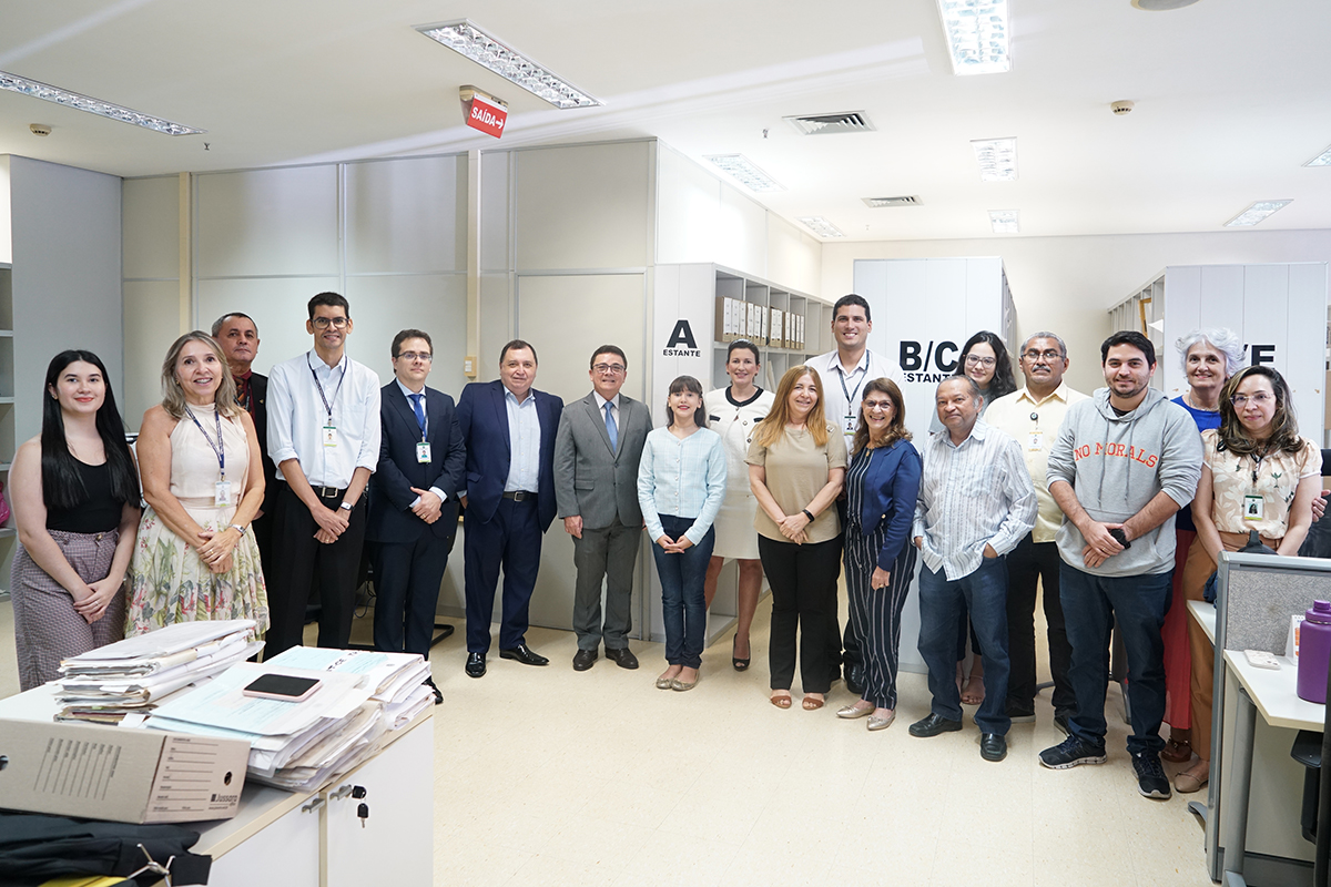 Em um ambiente de escritório, um grupo de dezoito pessoas está posando para uma foto. Homens e mulheres de diferentes idades compõem o grupo, vestidos de forma variada entre casual e social. Há pilhas de documentos e objetos de escritório visíveis em primeiro plano e ao fundo, com um letreiro de "SAÍDA" e placas de "ESTANTE" penduradas nas paredes.