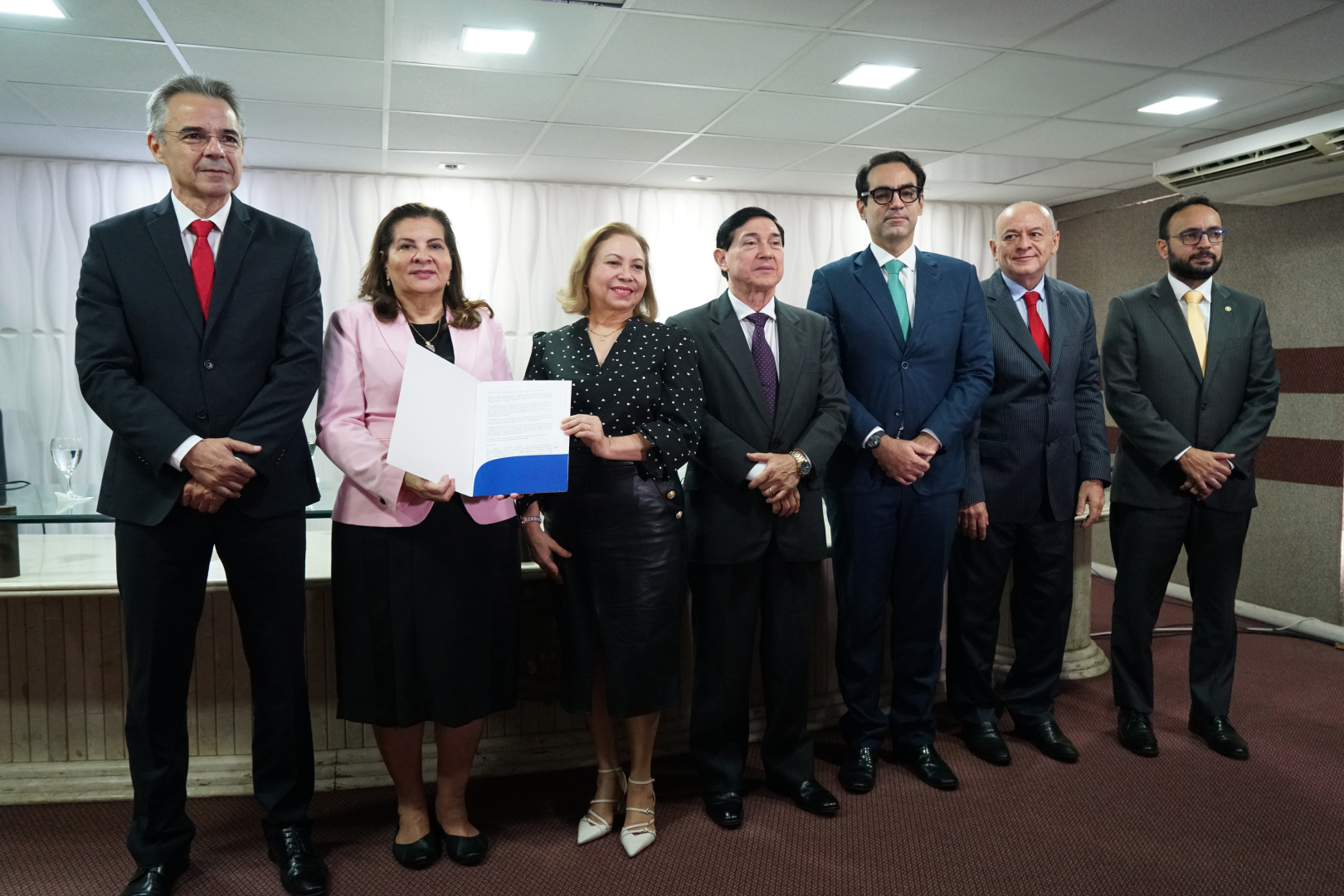 Sete pessoas, incluindo uma mulher no centro segurando um documento, posam em fila. Todos vestem trajes formais em um ambiente interno bem iluminado. A imagem captura um momento de formalidade e colaboração.